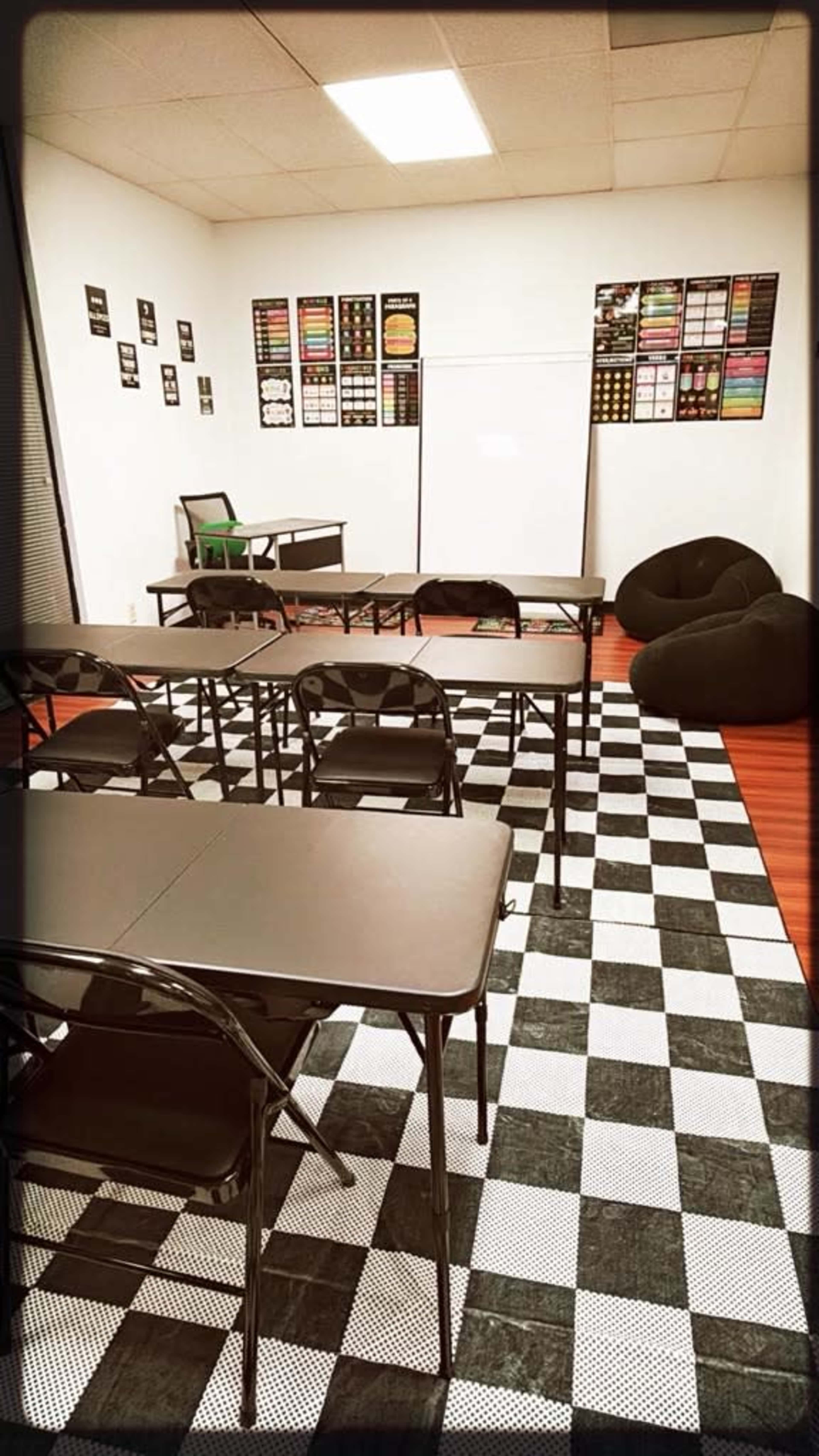 The image shows a classroom featuring several black folding tables and chairs arranged on a checkered floor, with a blank whiteboard and colorful posters on the walls.