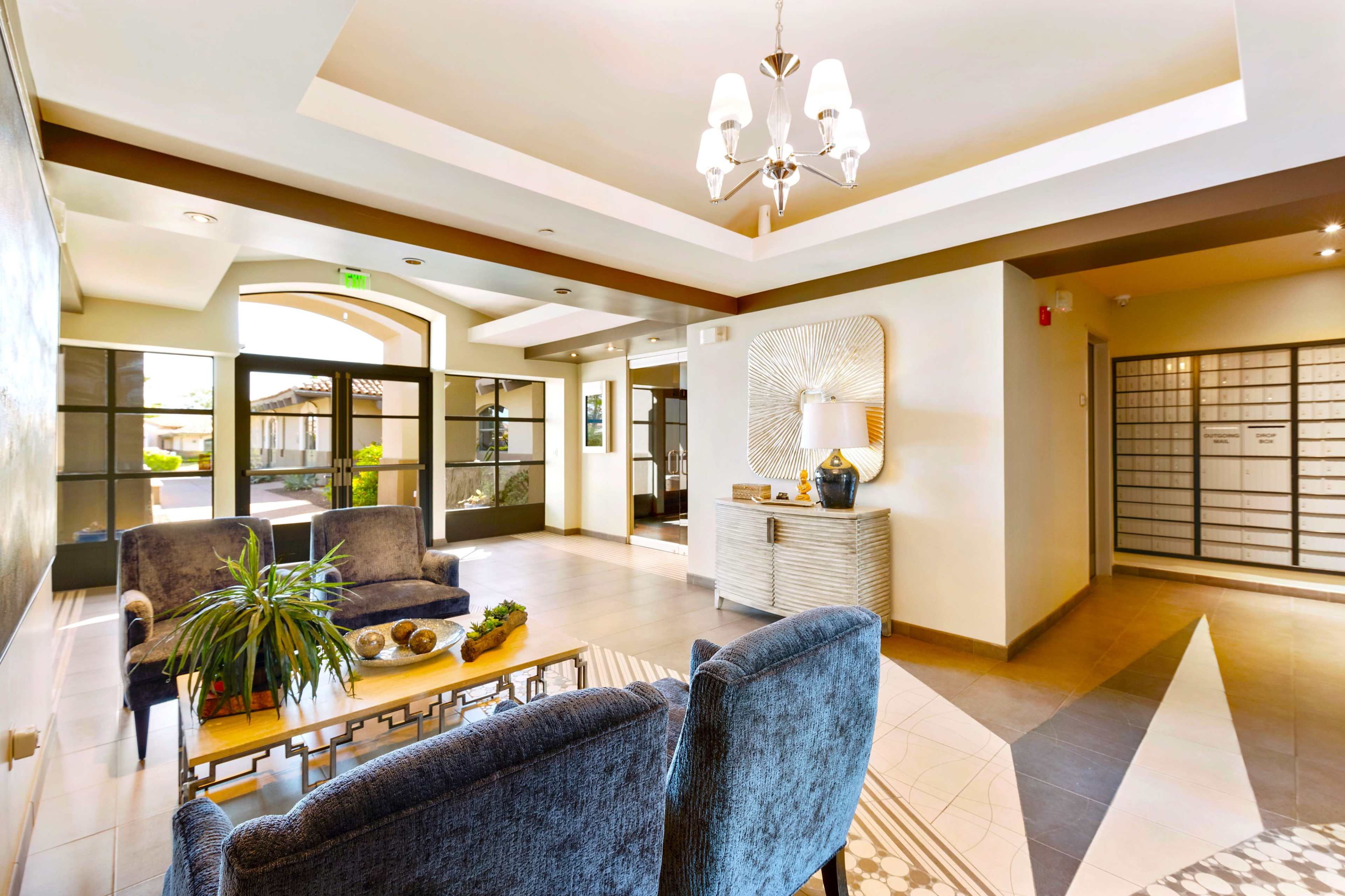 The image shows a spacious lobby area with two upholstered chairs, a decorative table, and a wall of mailboxes on one side.