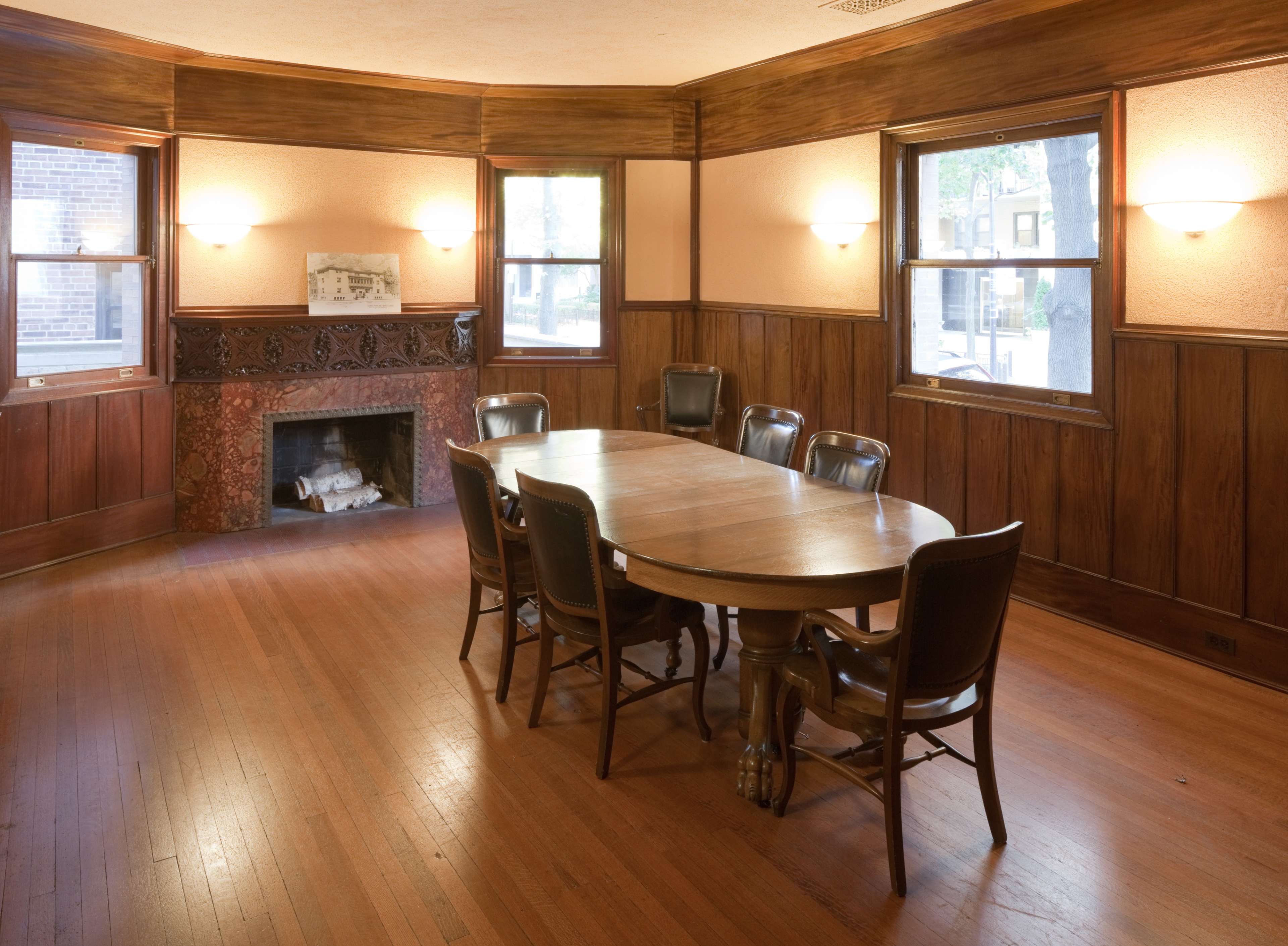 A wooden conference room features a large oval table surrounded by six leather chairs, with a fireplace and windows along the walls.