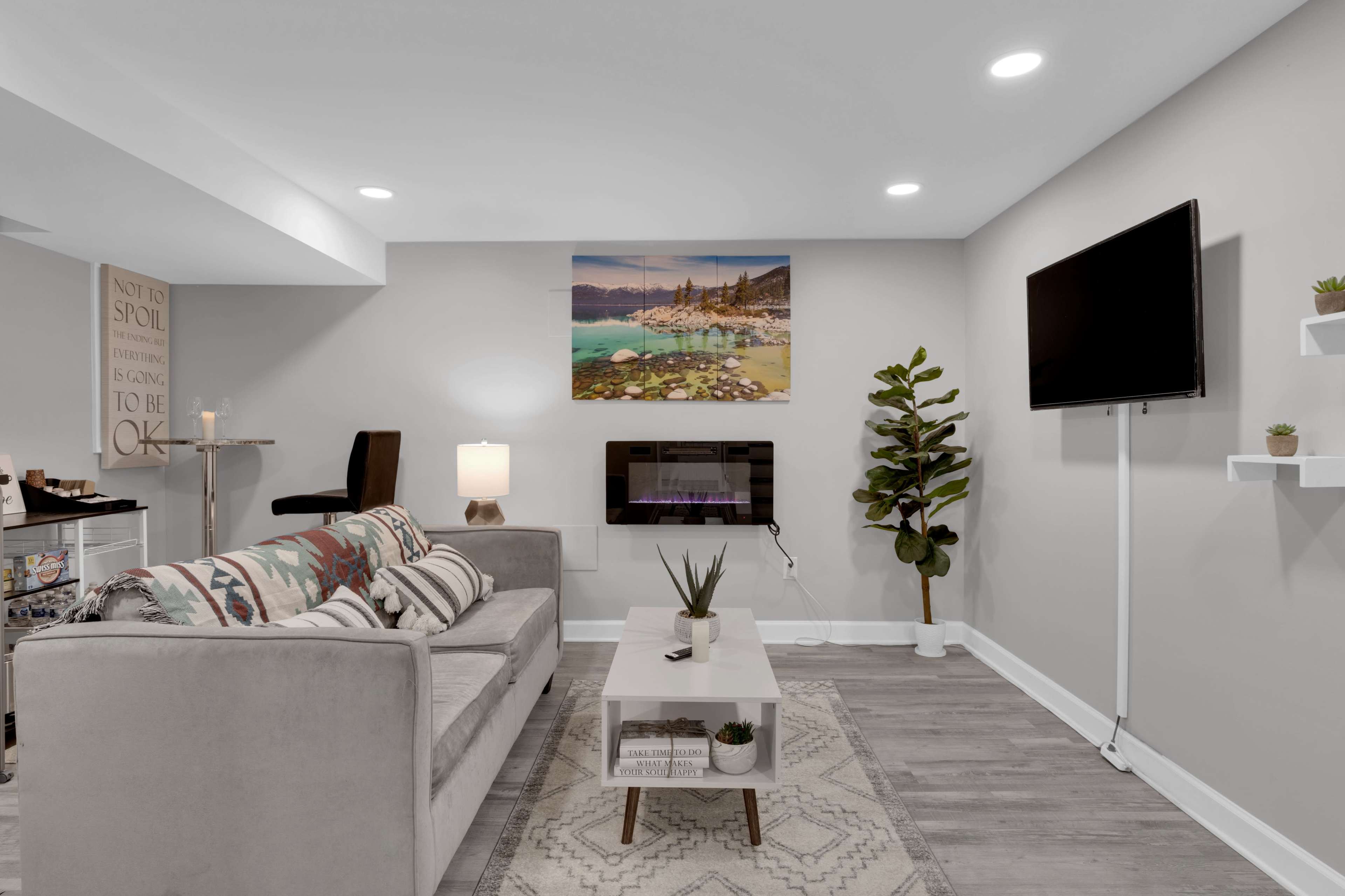 A modern living room with a light gray sofa, a wall-mounted TV, and a coffee table on a patterned rug, flanked by decorative plants.