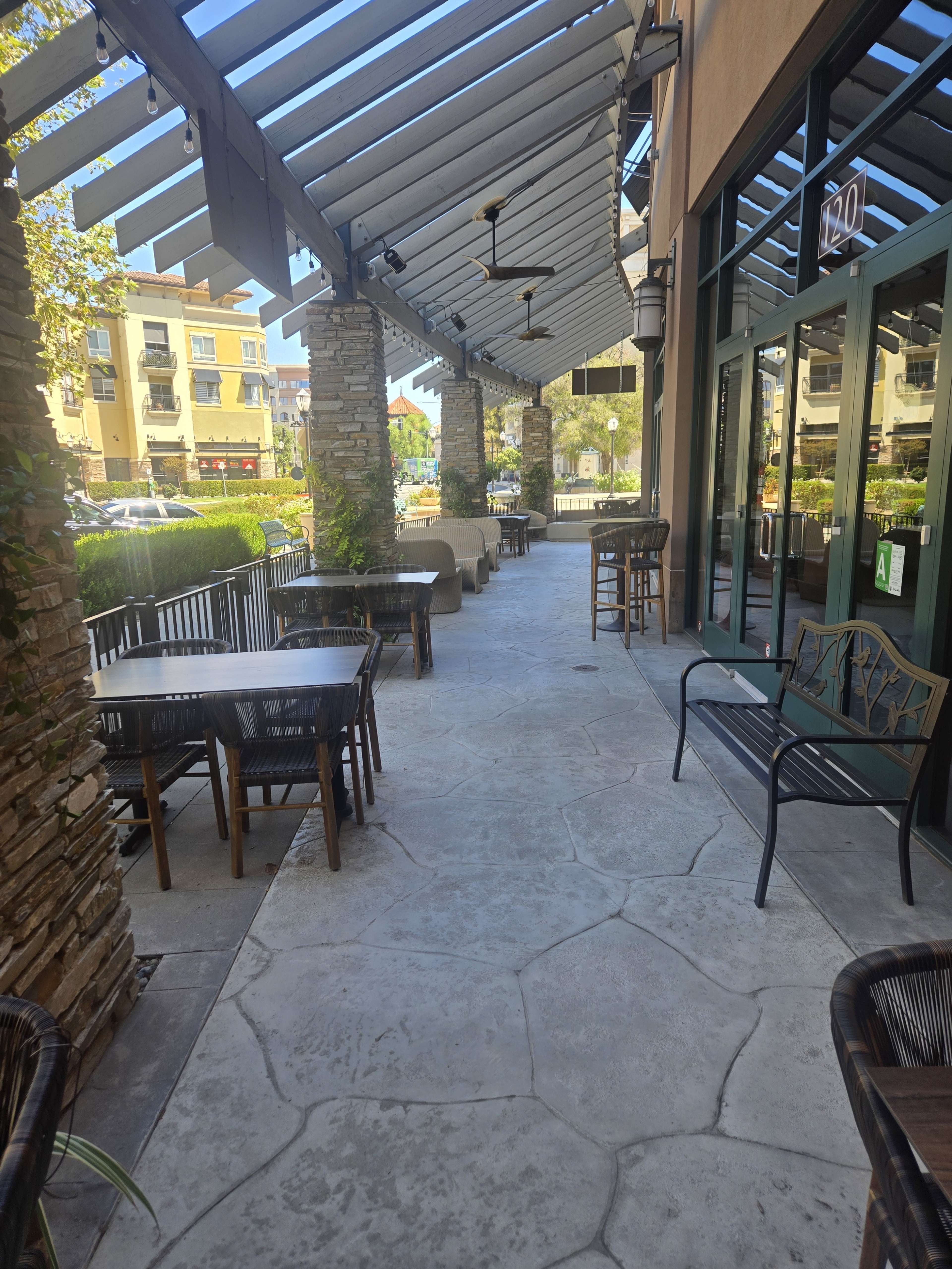 The image shows a long, shaded walkway with stone columns and outdoor seating at a restaurant or café.