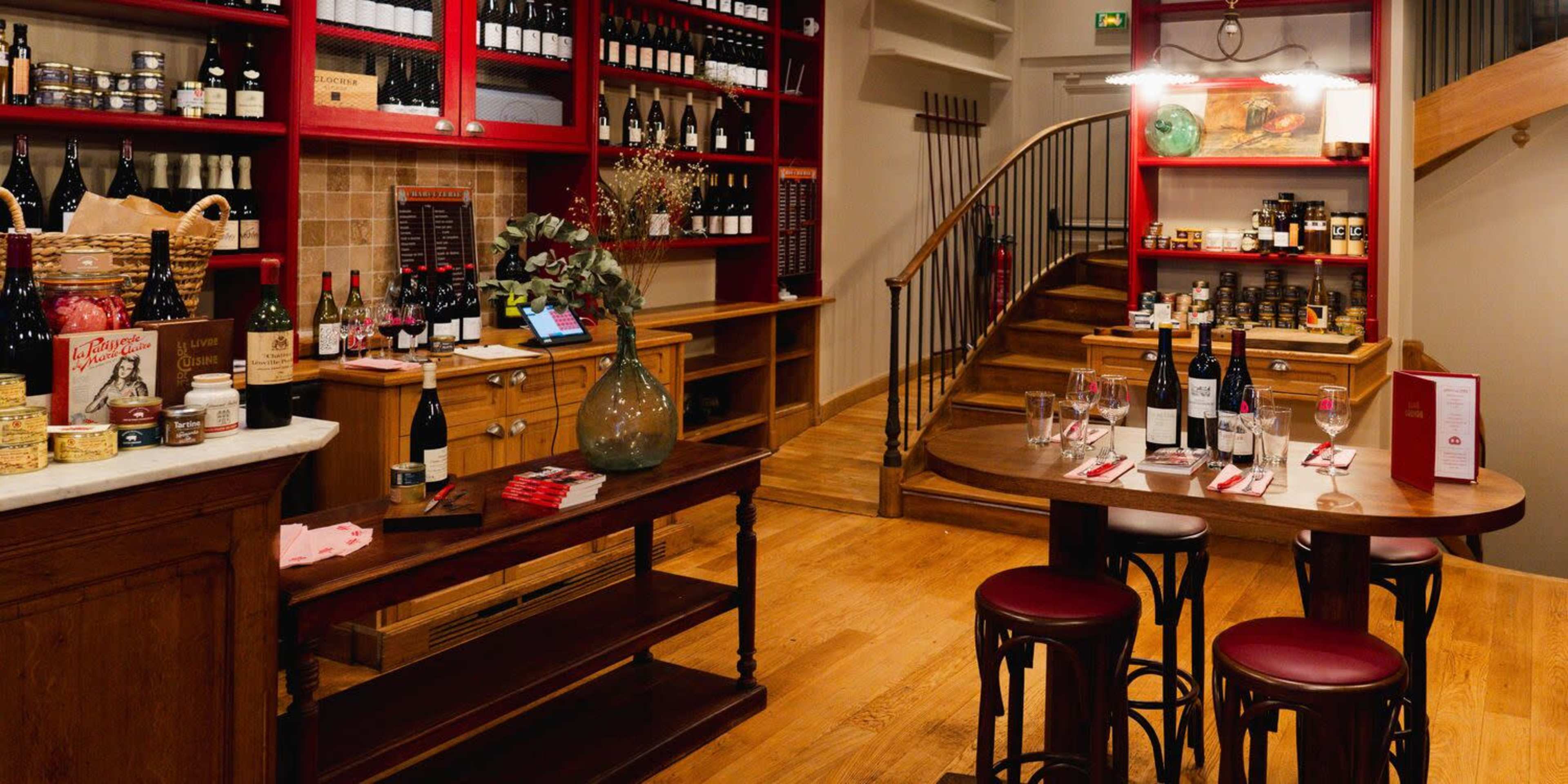 The image shows a wine shop interior featuring wooden shelves lined with bottles, a display of gourmet food items, and a set dining area with tables and chairs.