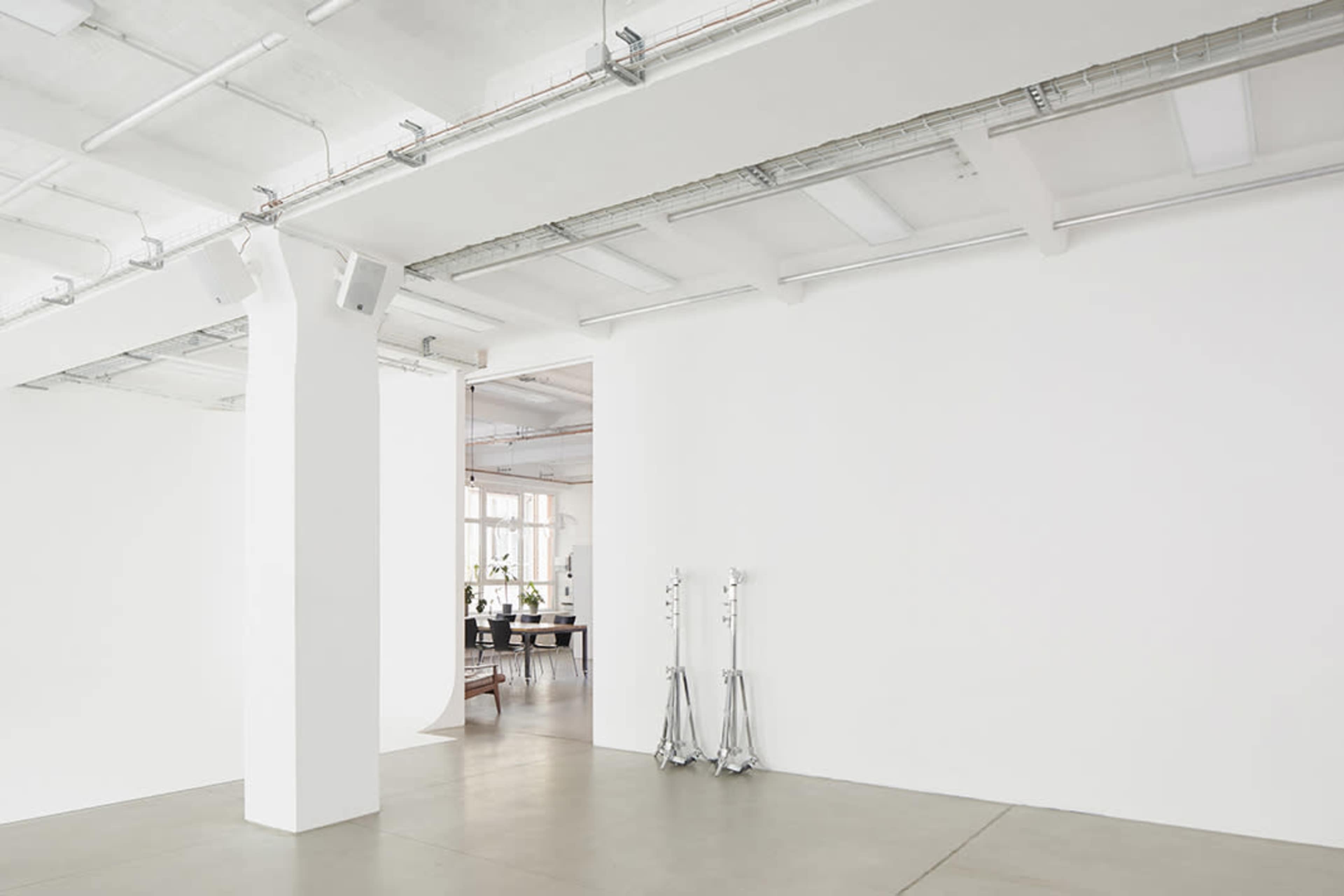 A minimalistic, well-lit studio space with white walls, concrete flooring, and lighting equipment along one side.