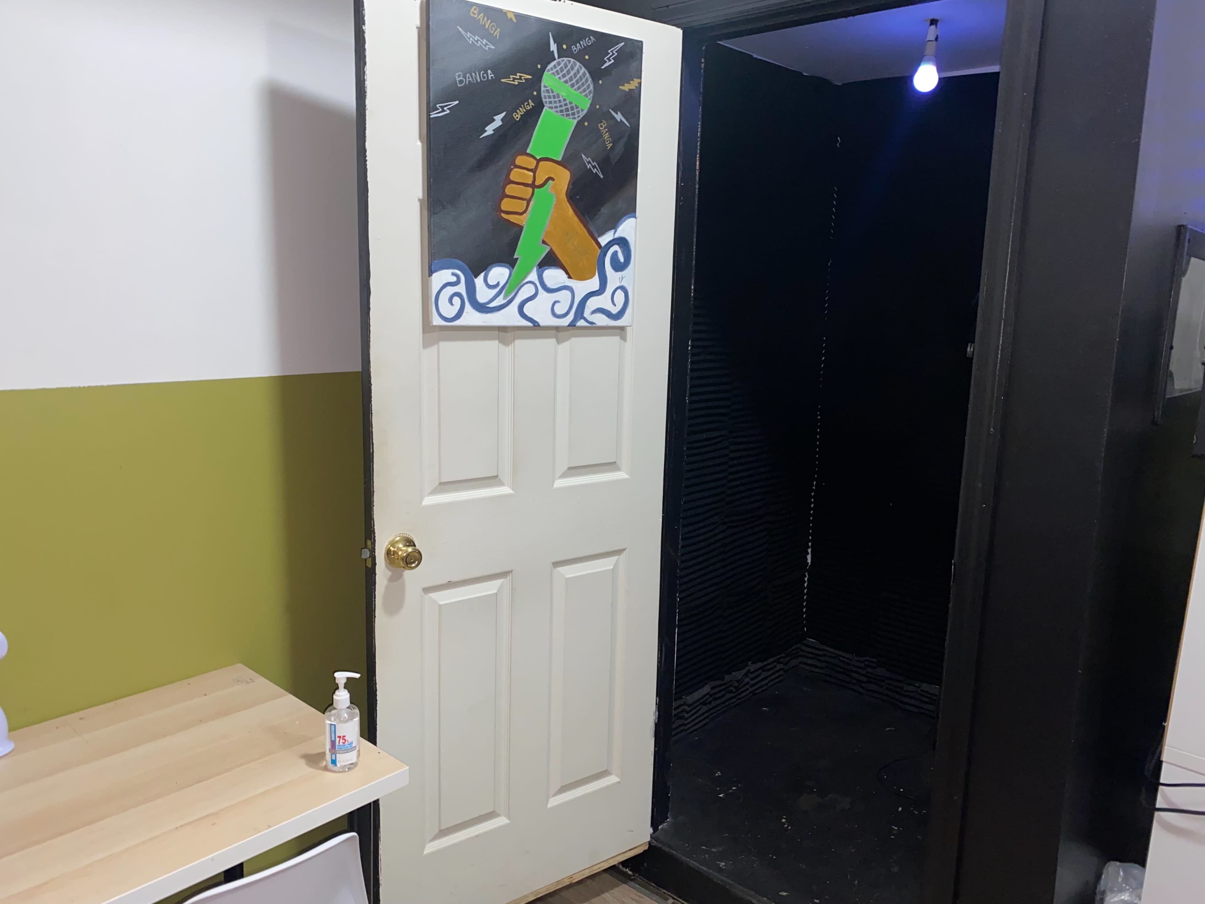 The image shows an open door leading to a soundproof booth with a microphone illustration on the door and a small table with hand sanitizer nearby.