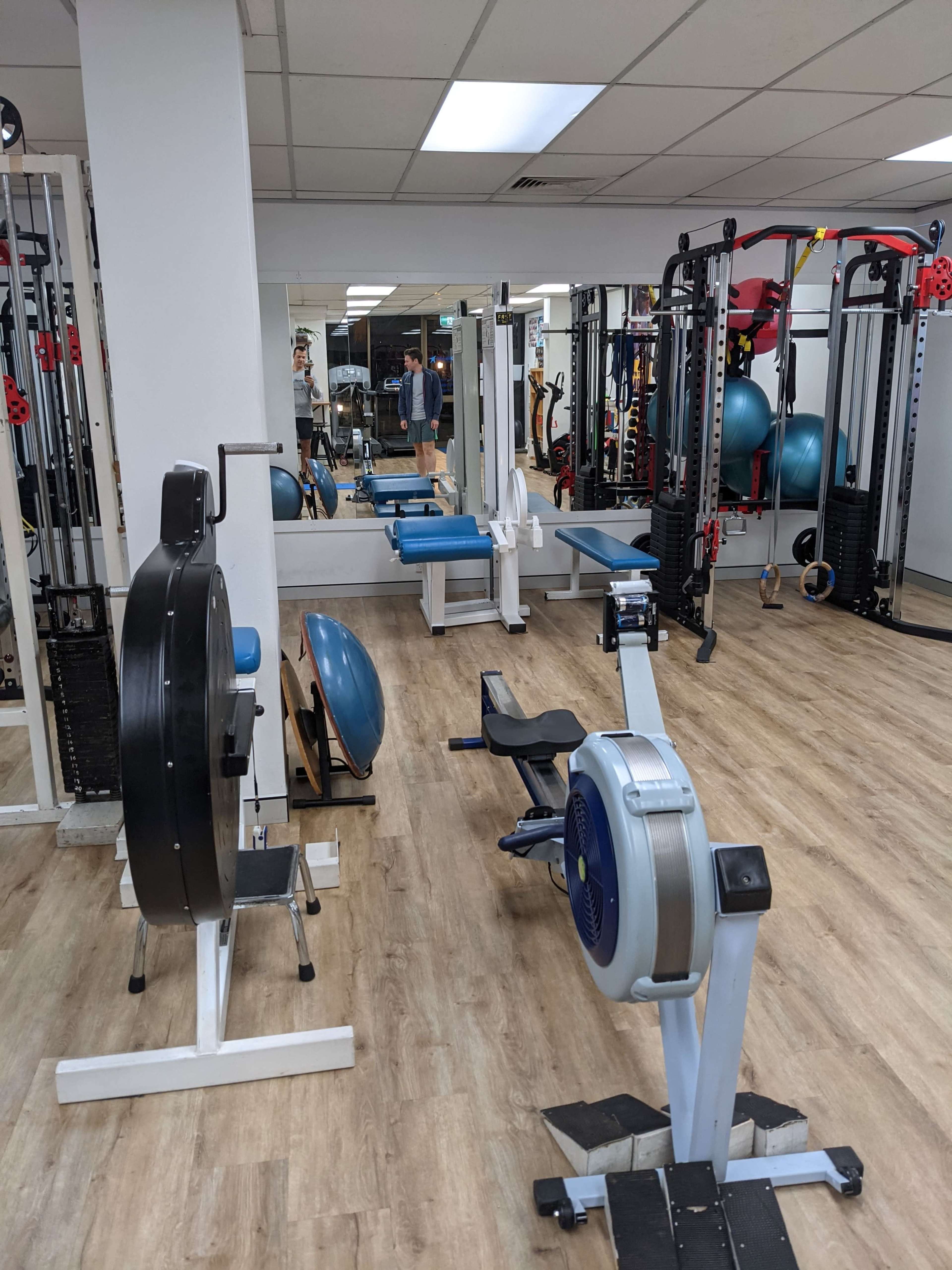 The image shows a fitness area with exercise equipment, including a rowing machine, free weights, and stability balls, set in a bright room with mirrors and wooden flooring.