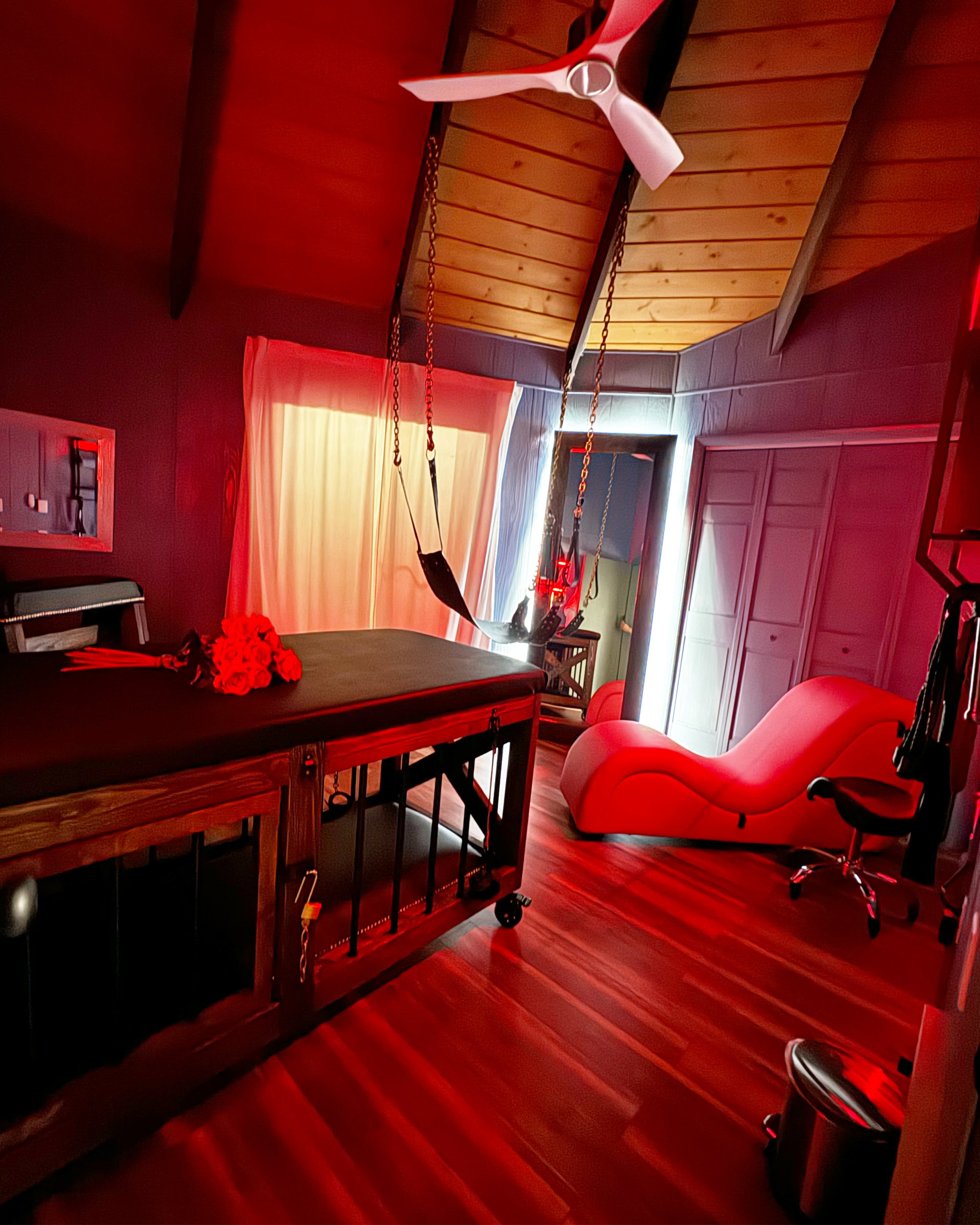 The room features a massage table, a hammock suspended from the ceiling, and a modern red chaise lounge, all under a wooden ceiling with a ceiling fan.