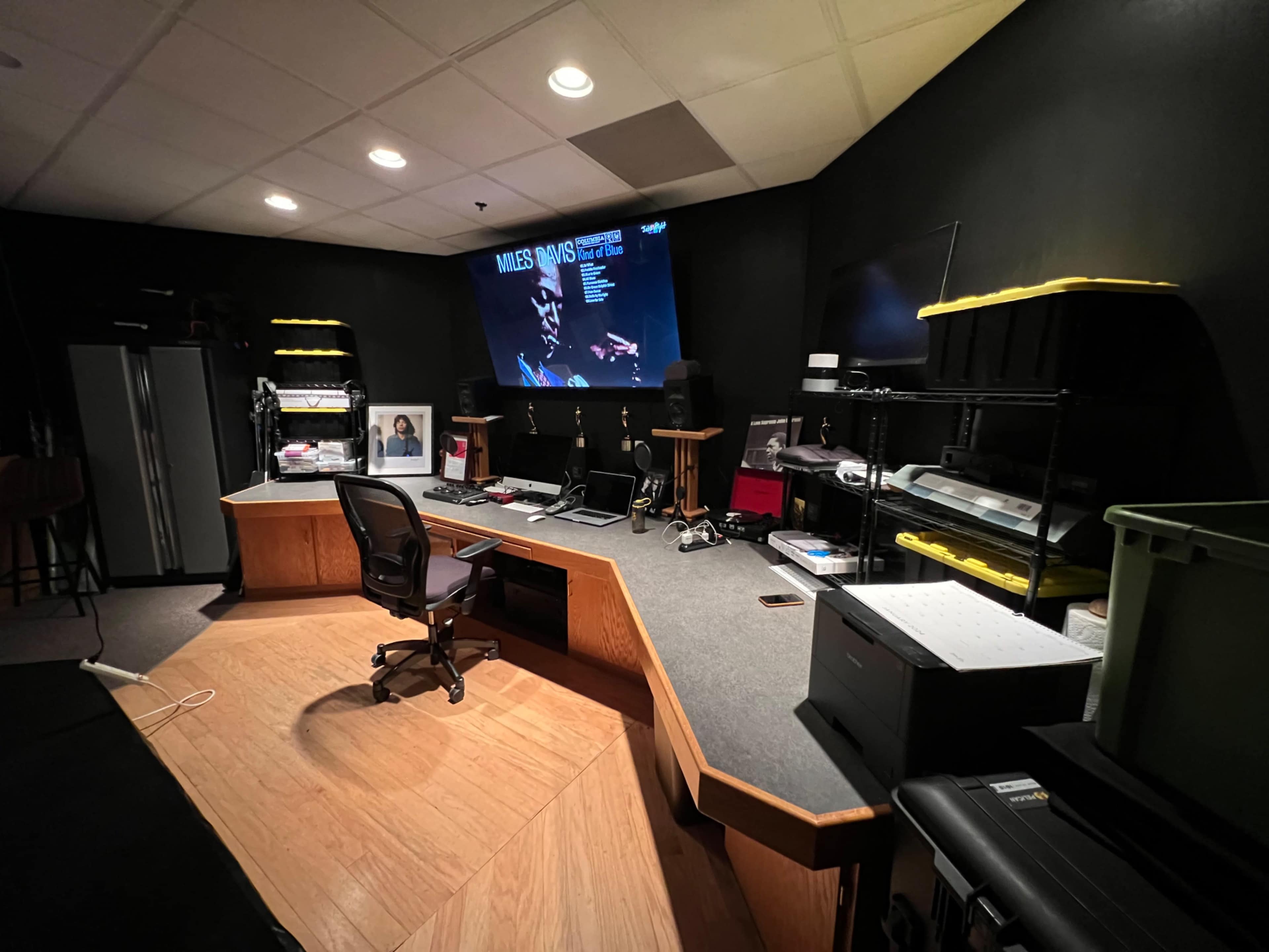 A spacious audio-visual production studio features a large screen displaying information, surrounded by various equipment, a desk with a chair, and storage shelves.