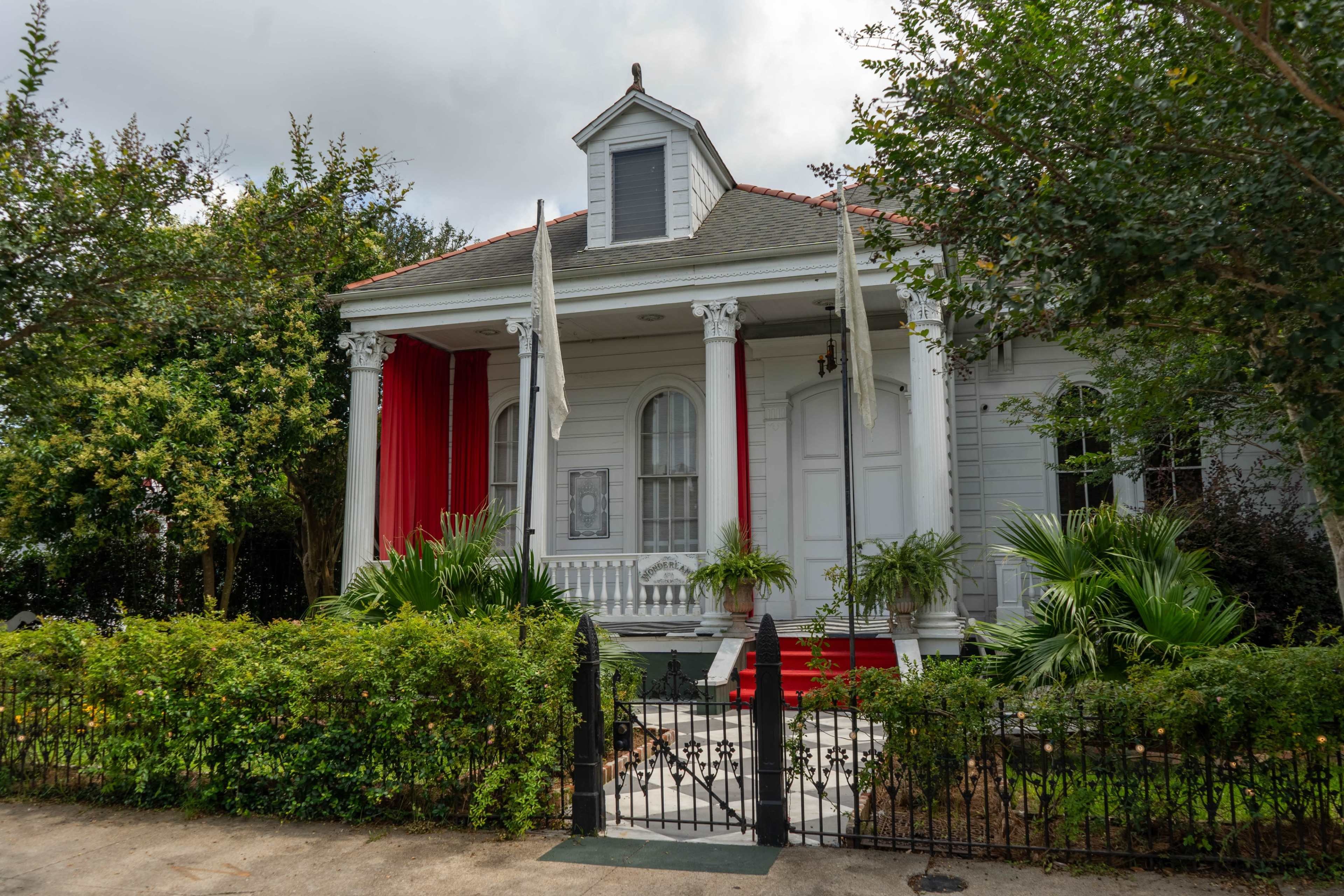A two-story house features a gray exterior with red accents, surrounded by greenery and a black wrought iron gate.