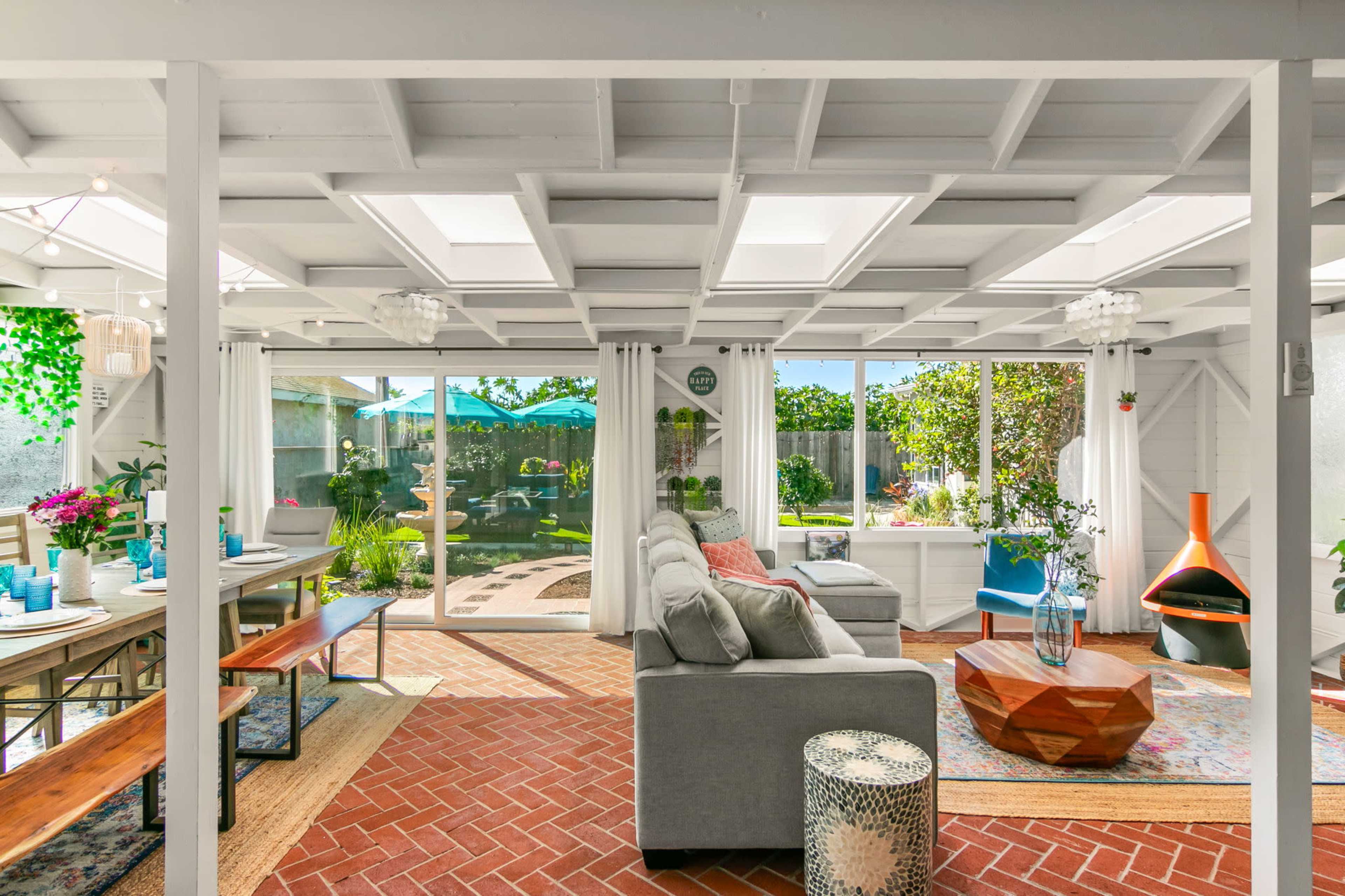 The image shows a bright, open living space with a cozy seating area, a dining table, and large glass doors leading to a garden.
