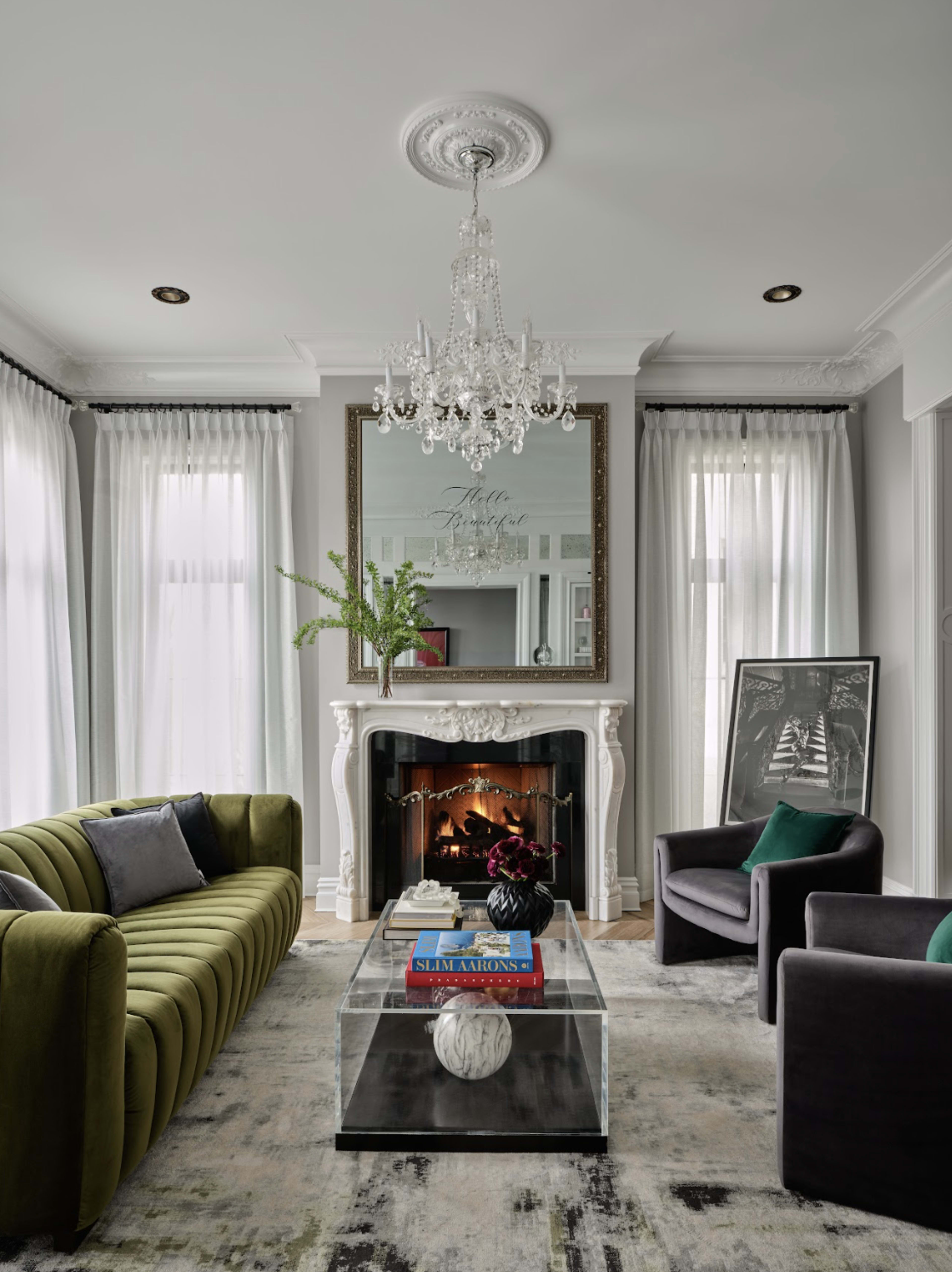 The living room features a green velvet sofa and armchairs, a glass coffee table, a fireplace with a white mantel, and a large mirror hanging above it.