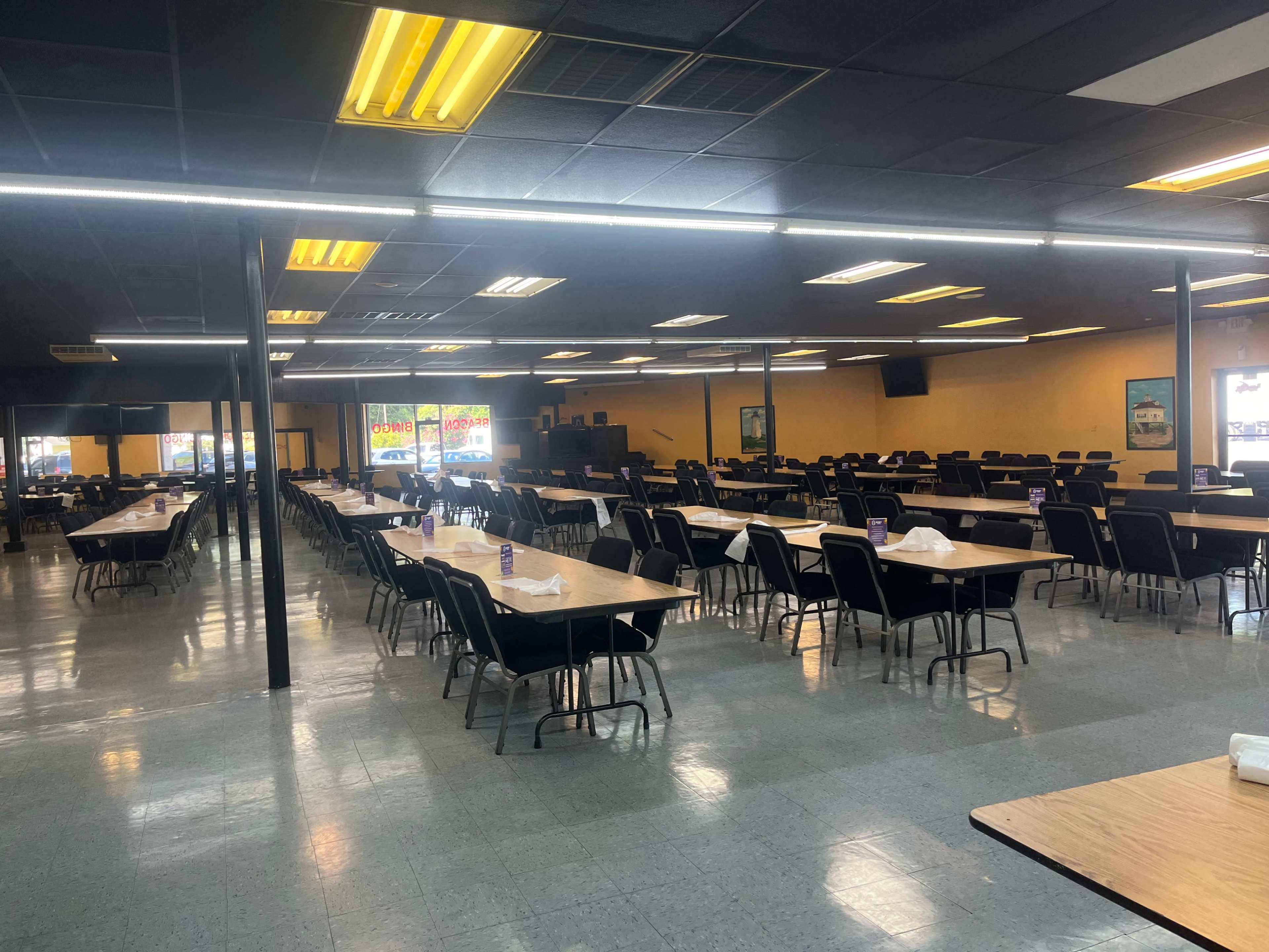 A large, empty dining area with multiple rows of tables and chairs, illuminated by overhead lights.