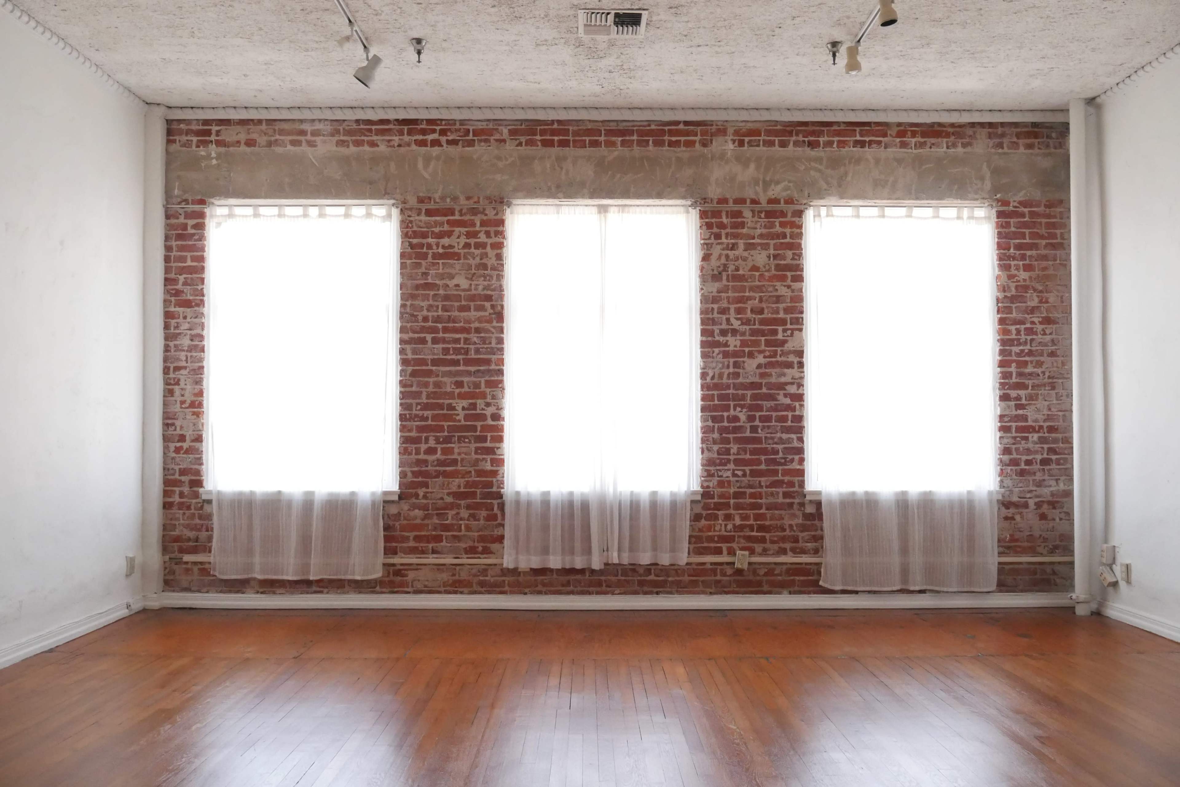 A room with three large windows covered by sheer curtains, and a wall of exposed red brick along with a wooden floor.
