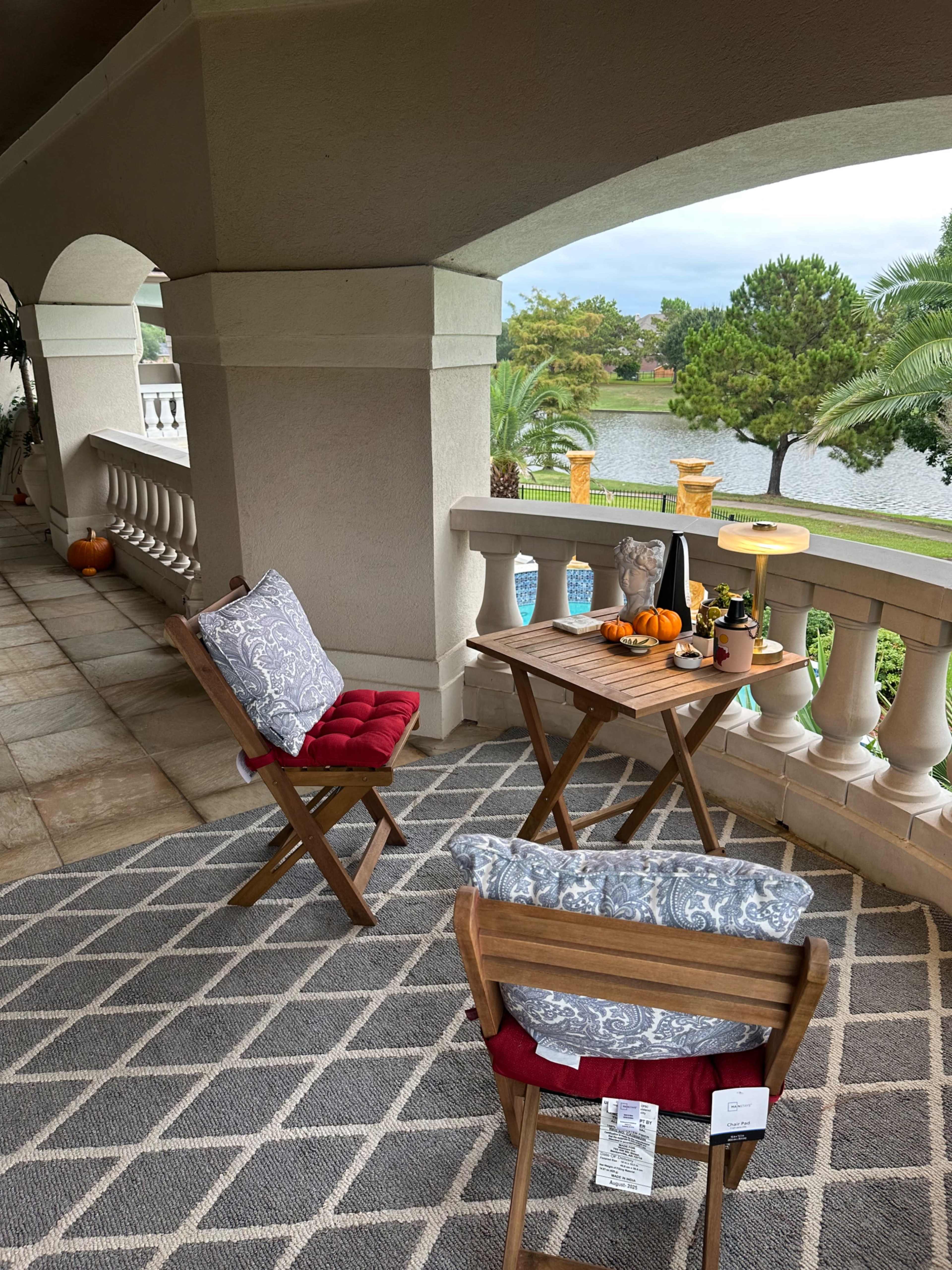 A cozy patio area features two folding chairs, a small wooden table with decorative items, and a view of a lake surrounded by greenery.