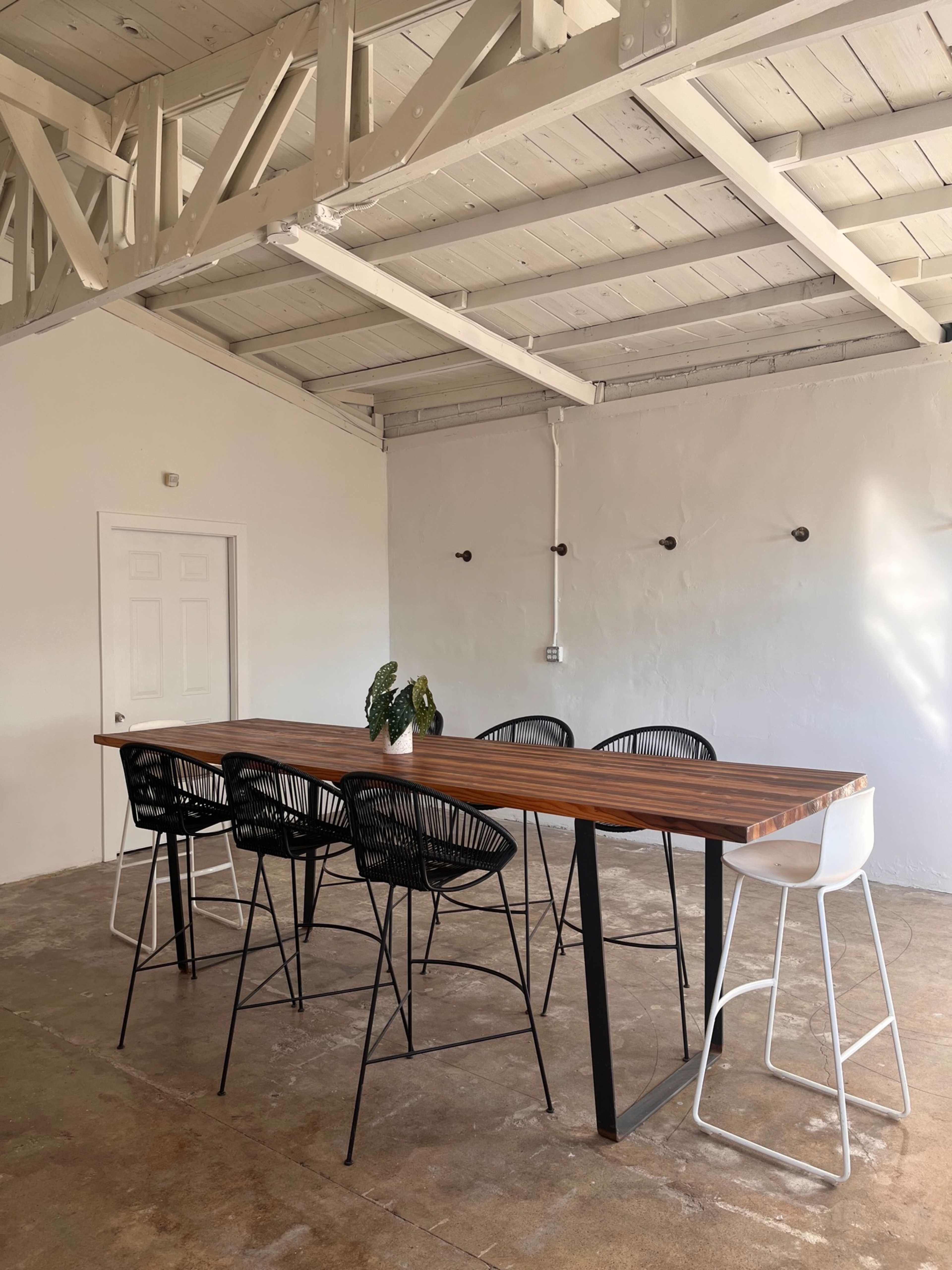A long wooden table with black and white high chairs is set in a minimalistic room with a white ceiling and walls.