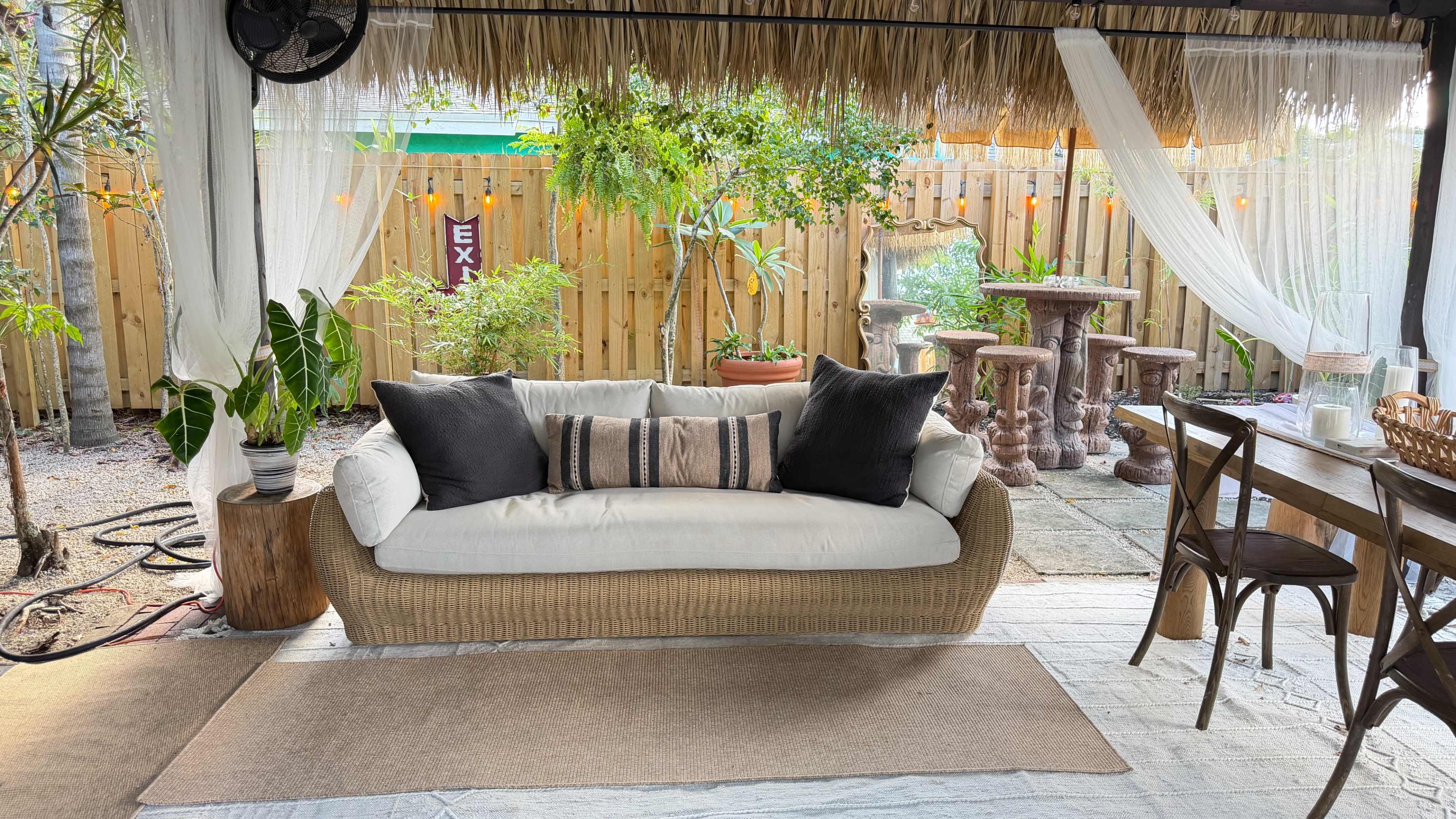 The image depicts a cozy outdoor seating area featuring a rattan sofa with cushions, surrounded by greenery and decorative elements.