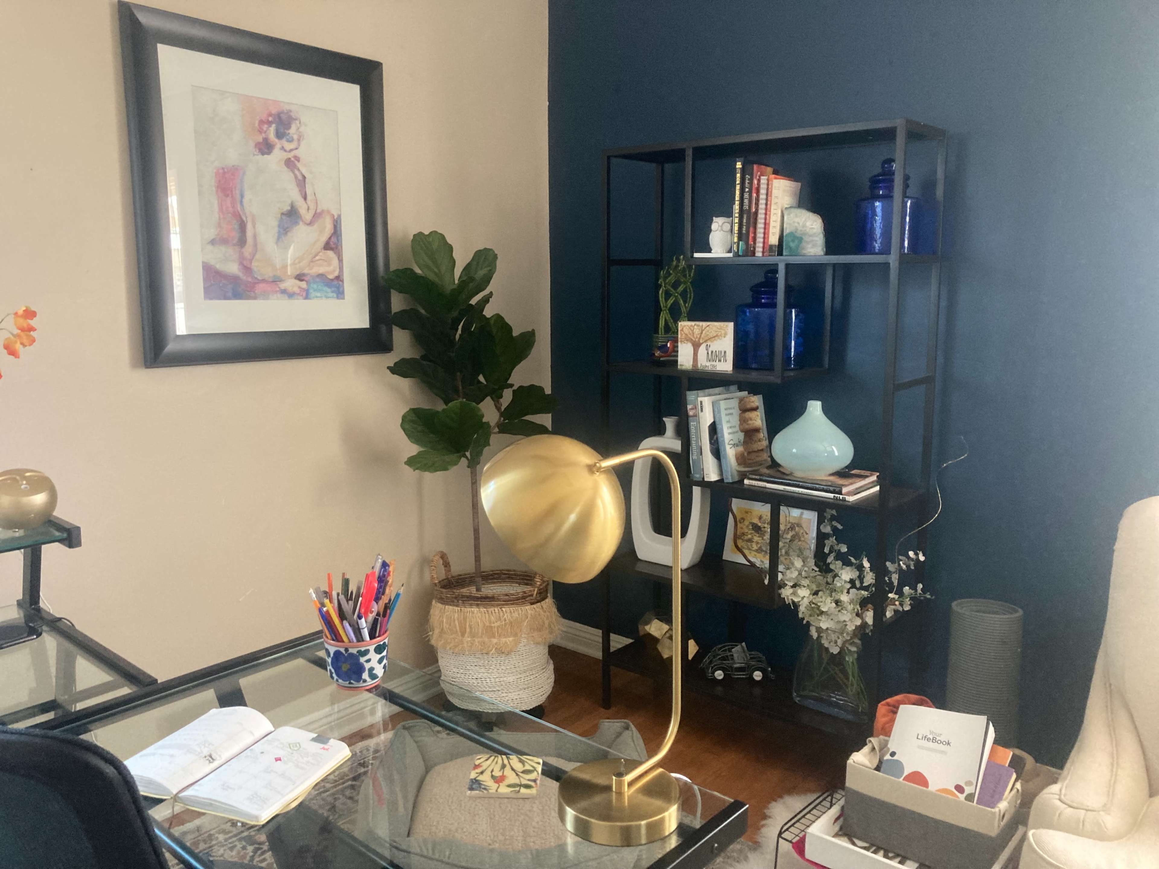A cozy office space featuring a glass desk with a gold lamp, a bookshelf filled with books and decorative items, and a potted plant.