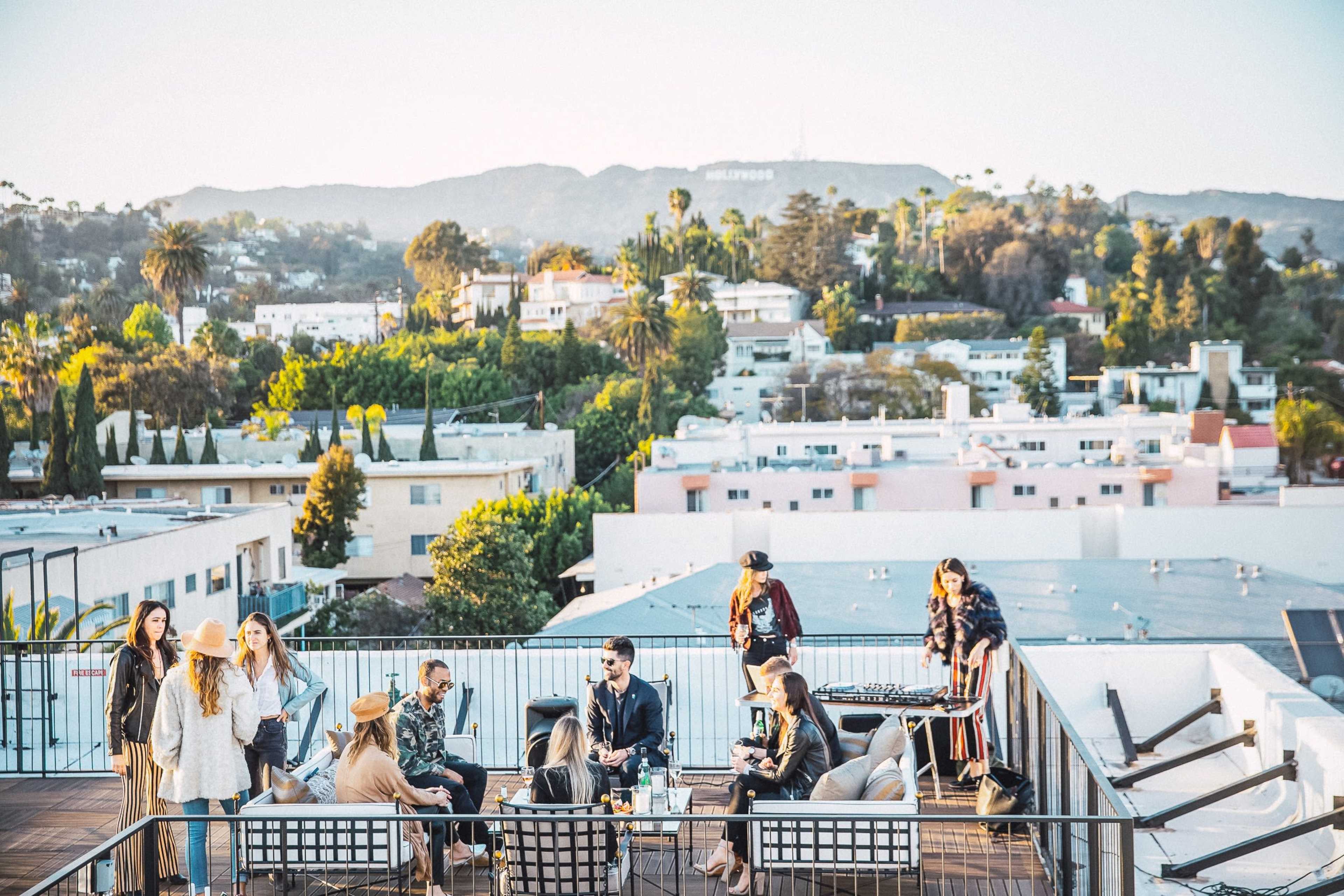 A group of people socializes on a rooftop deck overlooking a residential area with hills and trees in the background.