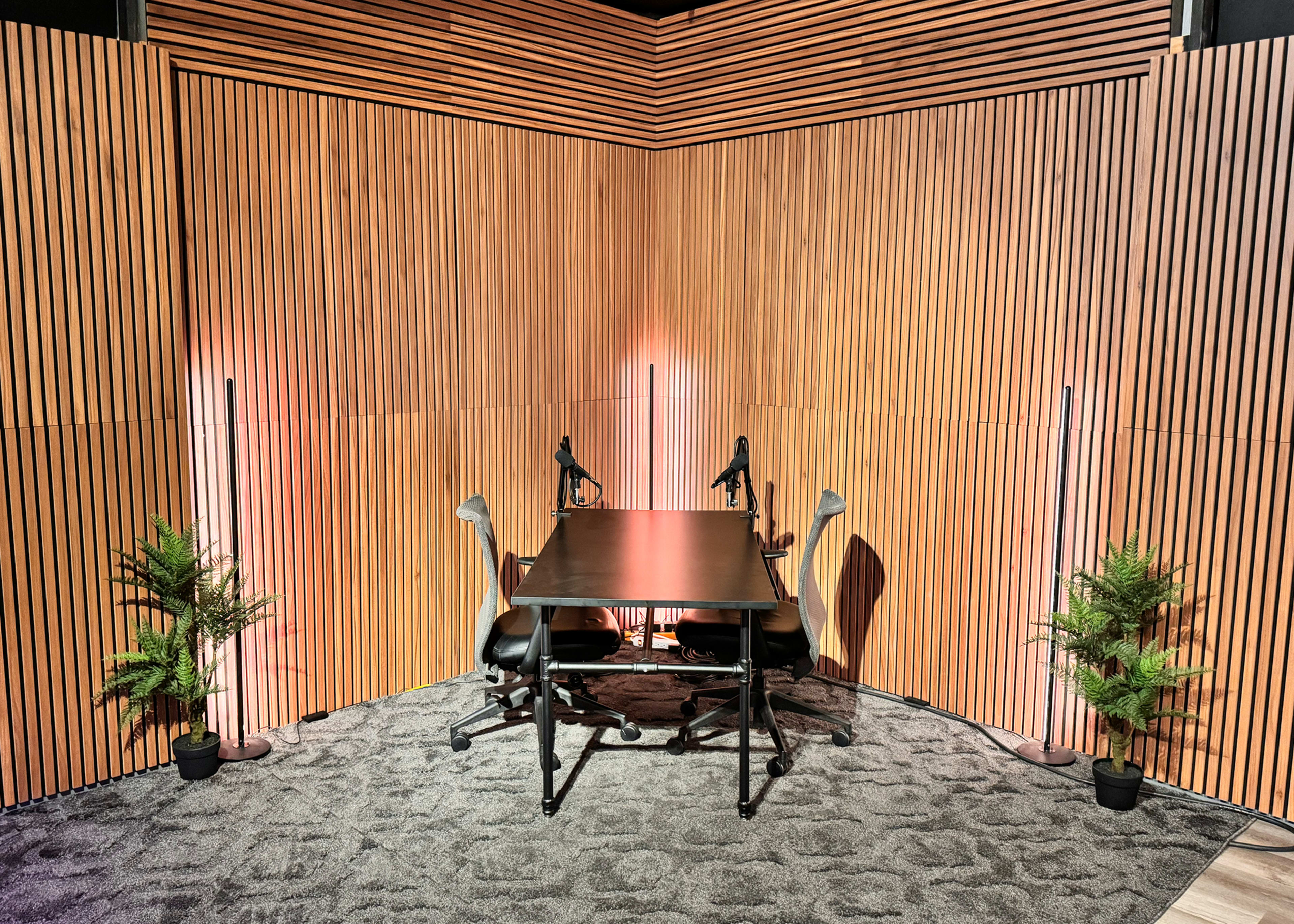 The image shows a modern corner studio setup with a black table and two chairs, surrounded by a wood-paneled wall and illuminated by soft lighting.