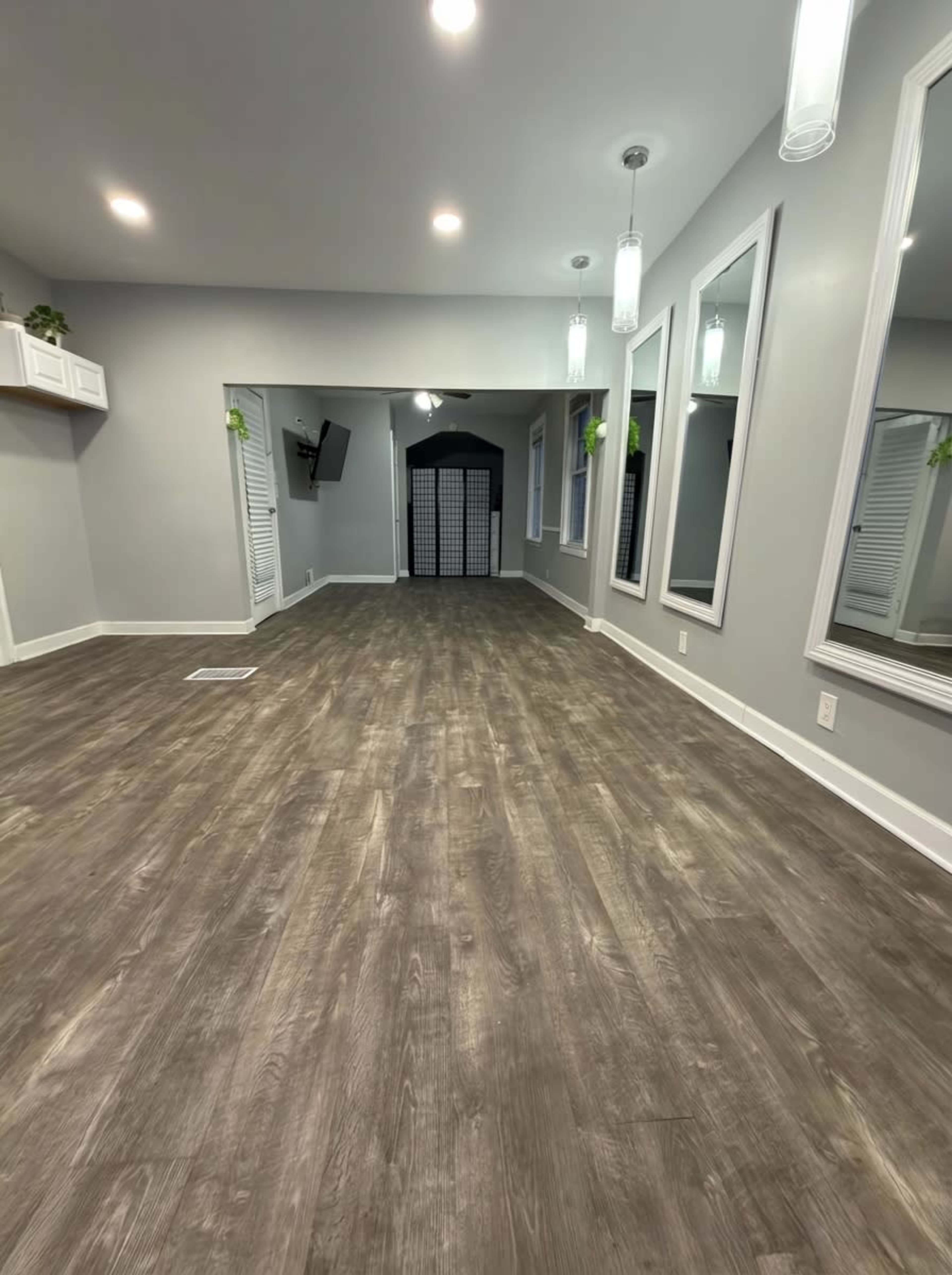 The image shows a spacious room with laminate flooring, two large wall mirrors, and minimal furniture, featuring bright lighting and a door leading to another area.