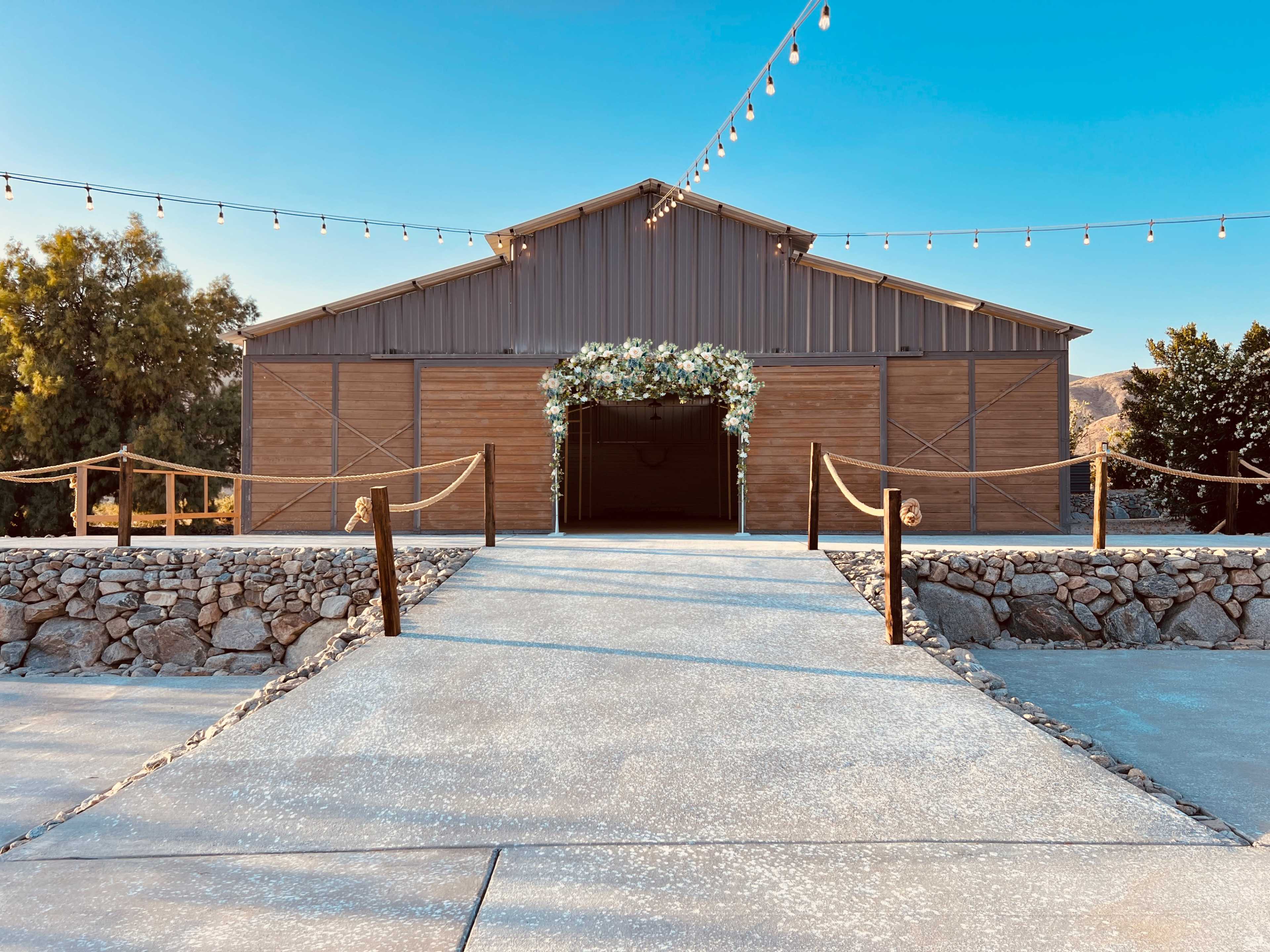 A large metal barn with wooden doors, decorated with flowers, set against a clear blue sky and connected by a gravel pathway.