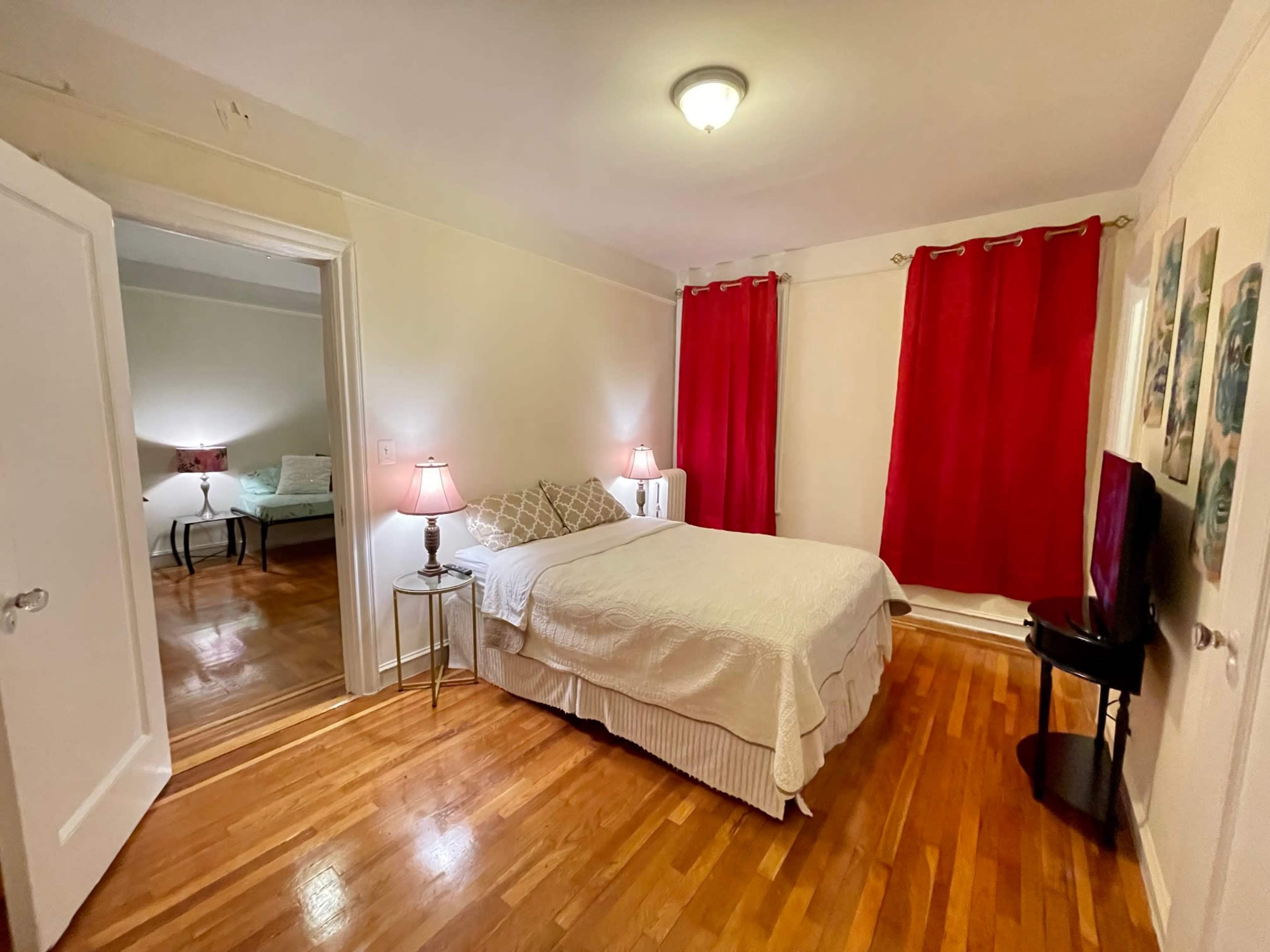 The image shows a comfortably furnished bedroom with a bed, nightstands, and red curtains, and an adjacent room is partially visible through an open doorway.