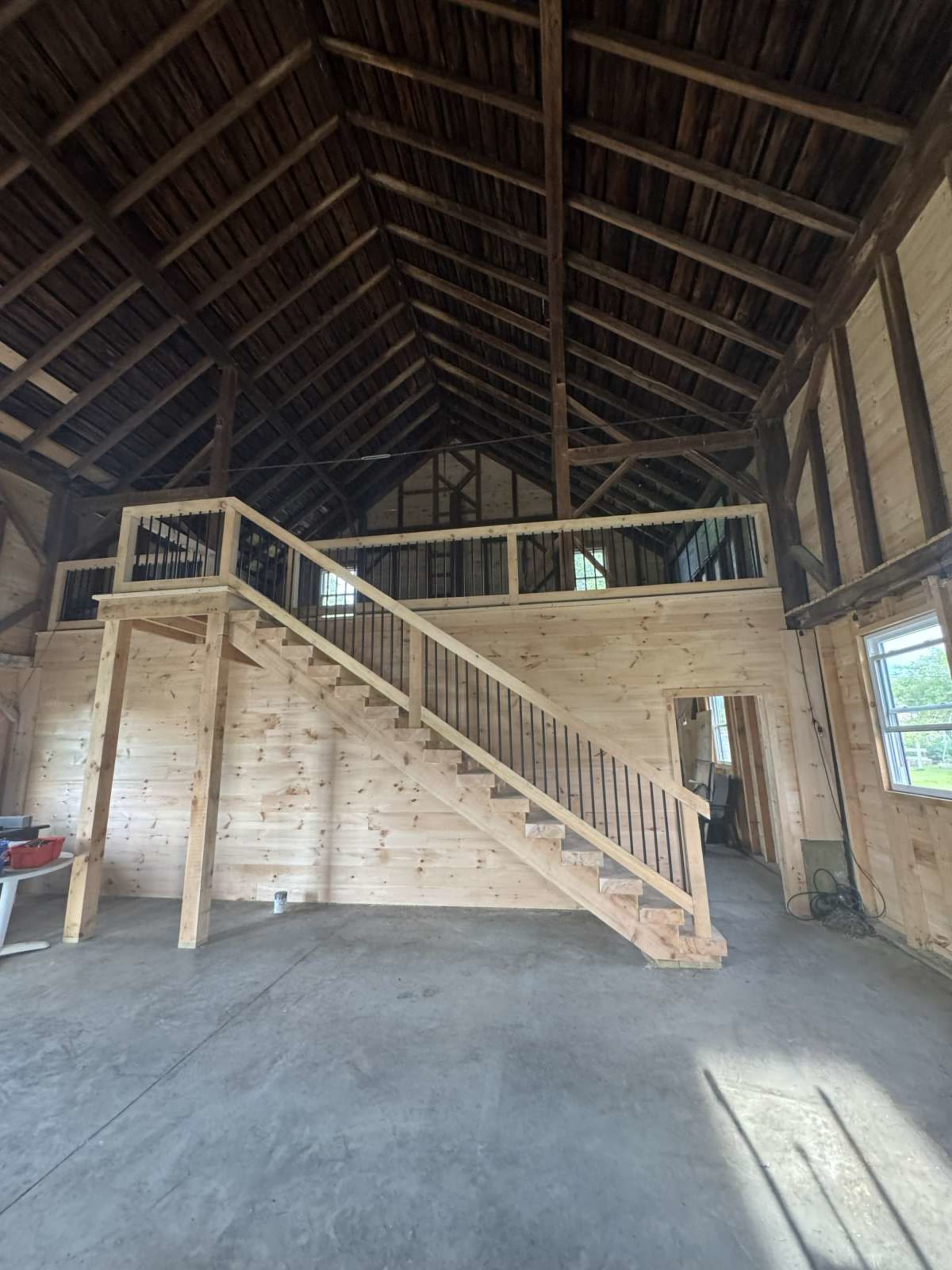 The image shows the interior of a wooden building with exposed beams, featuring a staircase leading to a lofted area.
