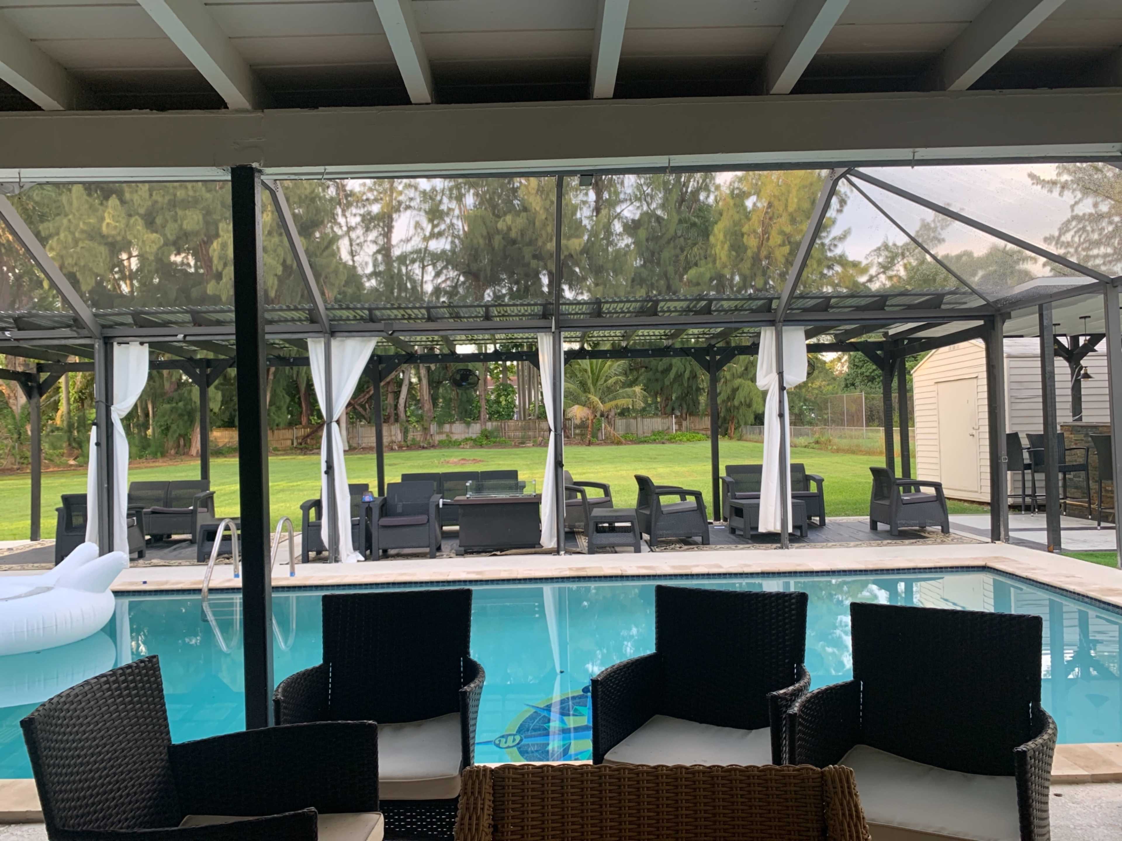 A covered patio with black seating surrounds a swimming pool, overlooking a lawn with trees in the background.