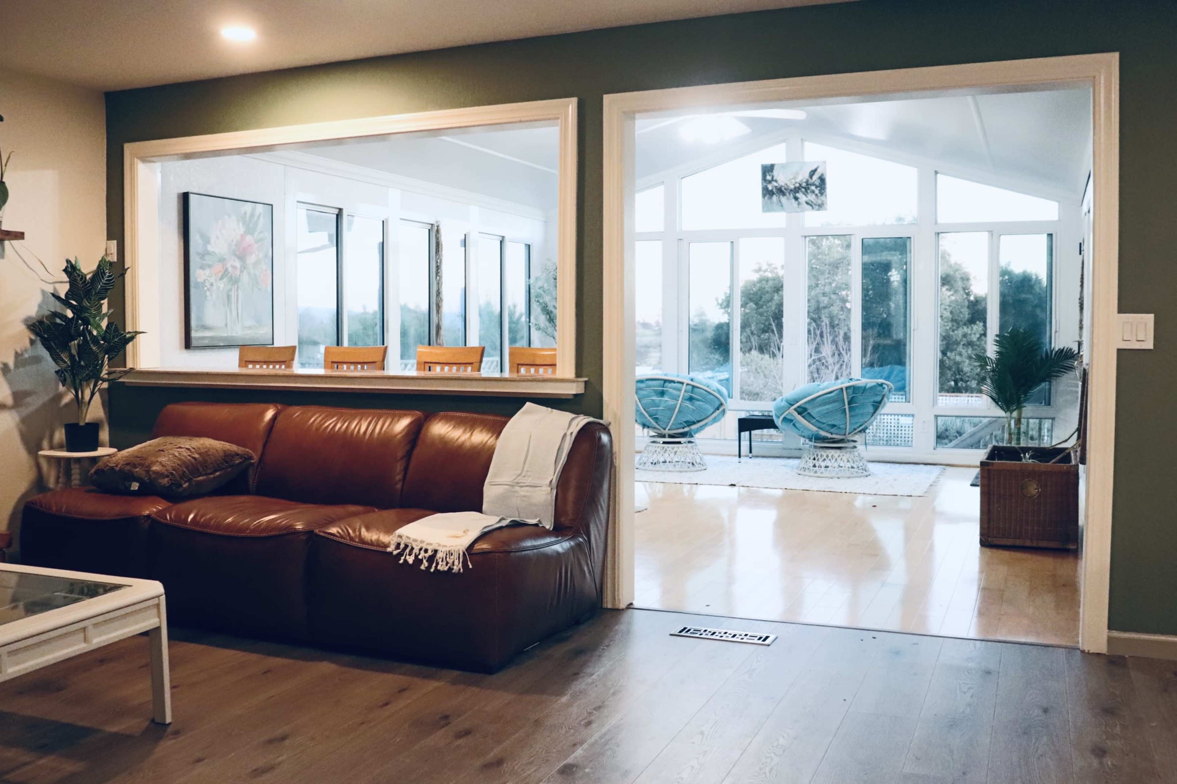 A leather couch faces a bright sunroom with large windows and wicker chairs.