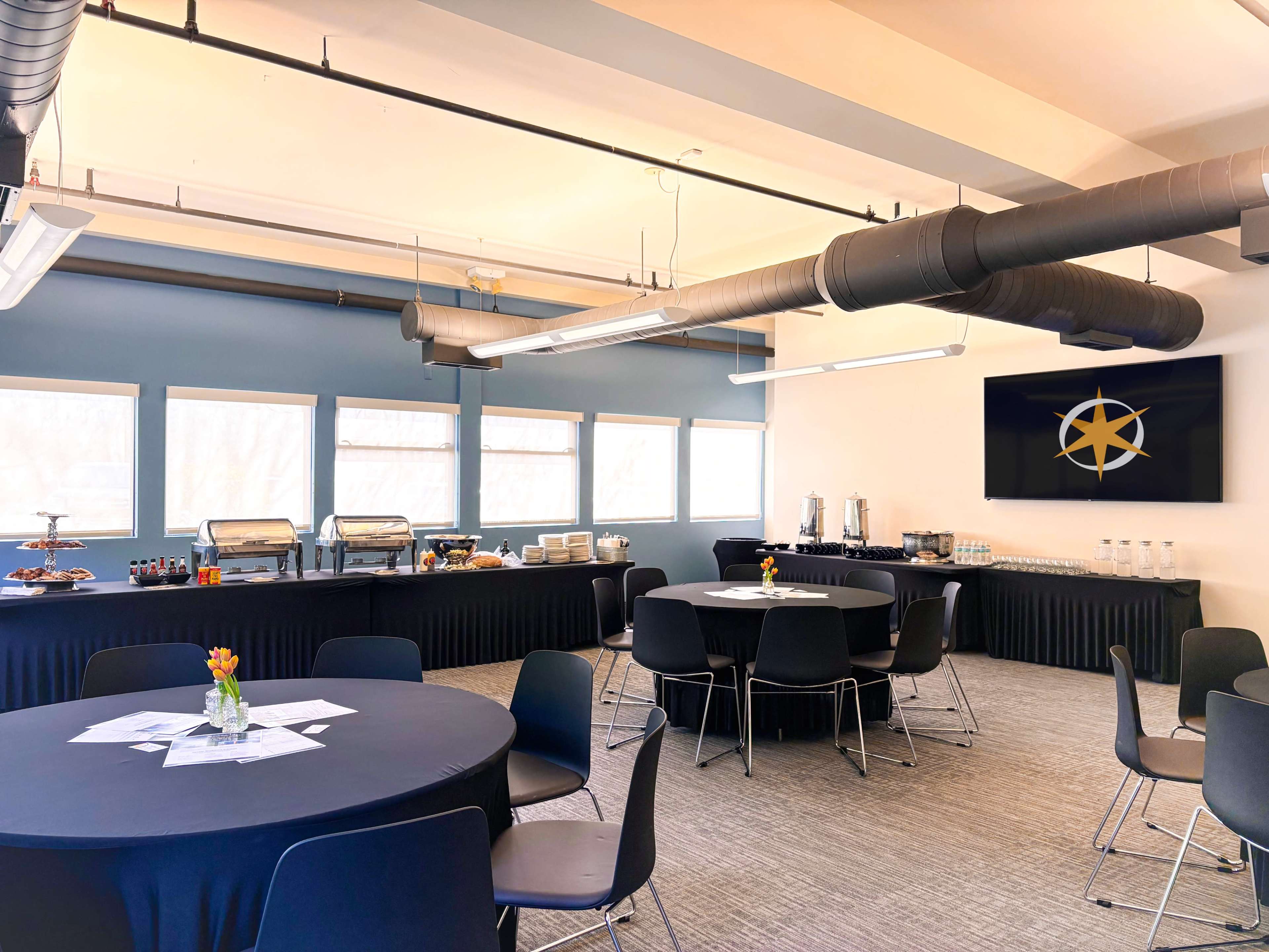A modern conference room features black circular tables and a buffet setup against a wall with a large screen displaying a logo.