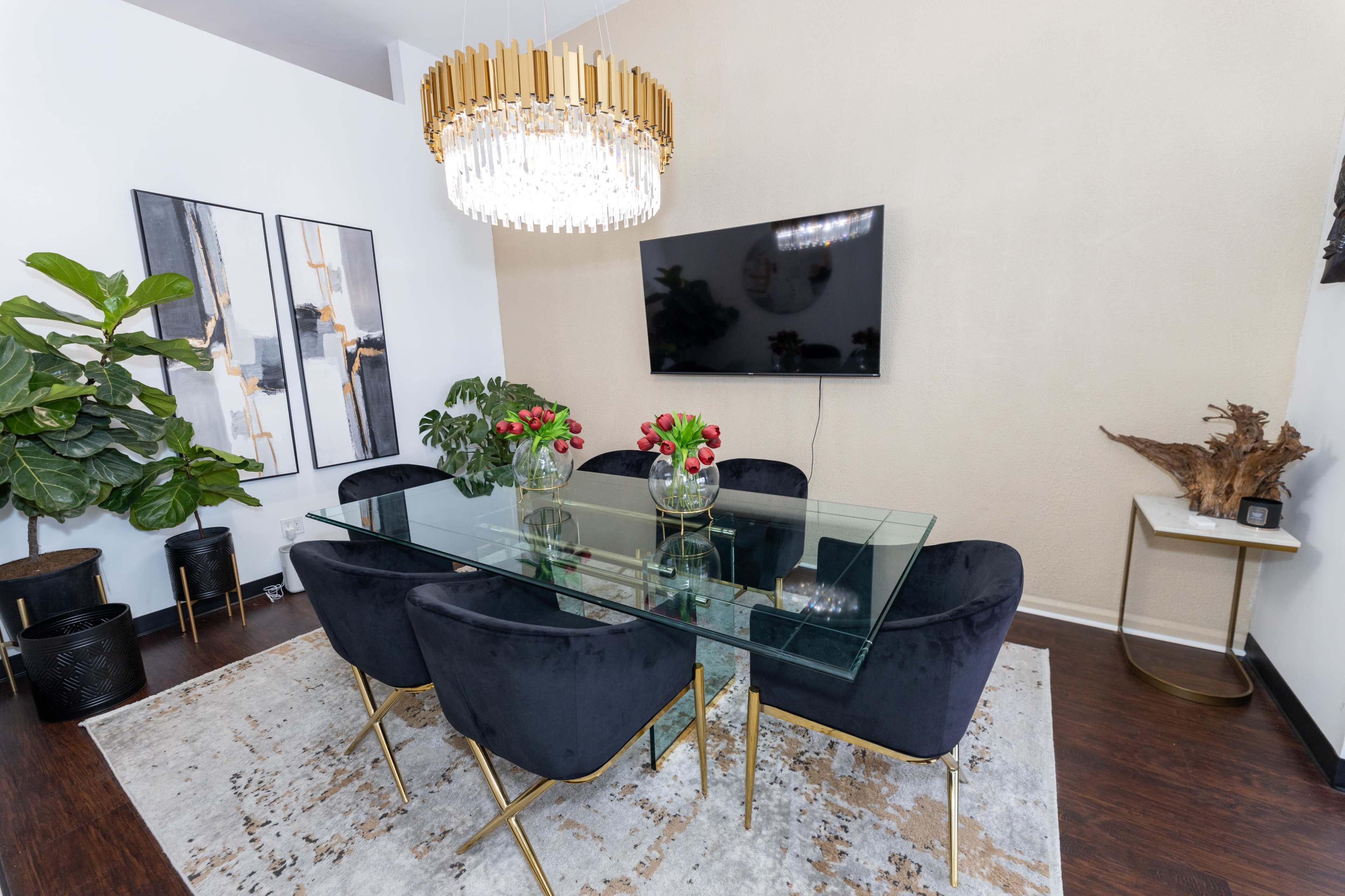 A modern dining room featuring a glass table surrounded by black chairs, a large chandelier overhead, and decorative plants along with wall art.