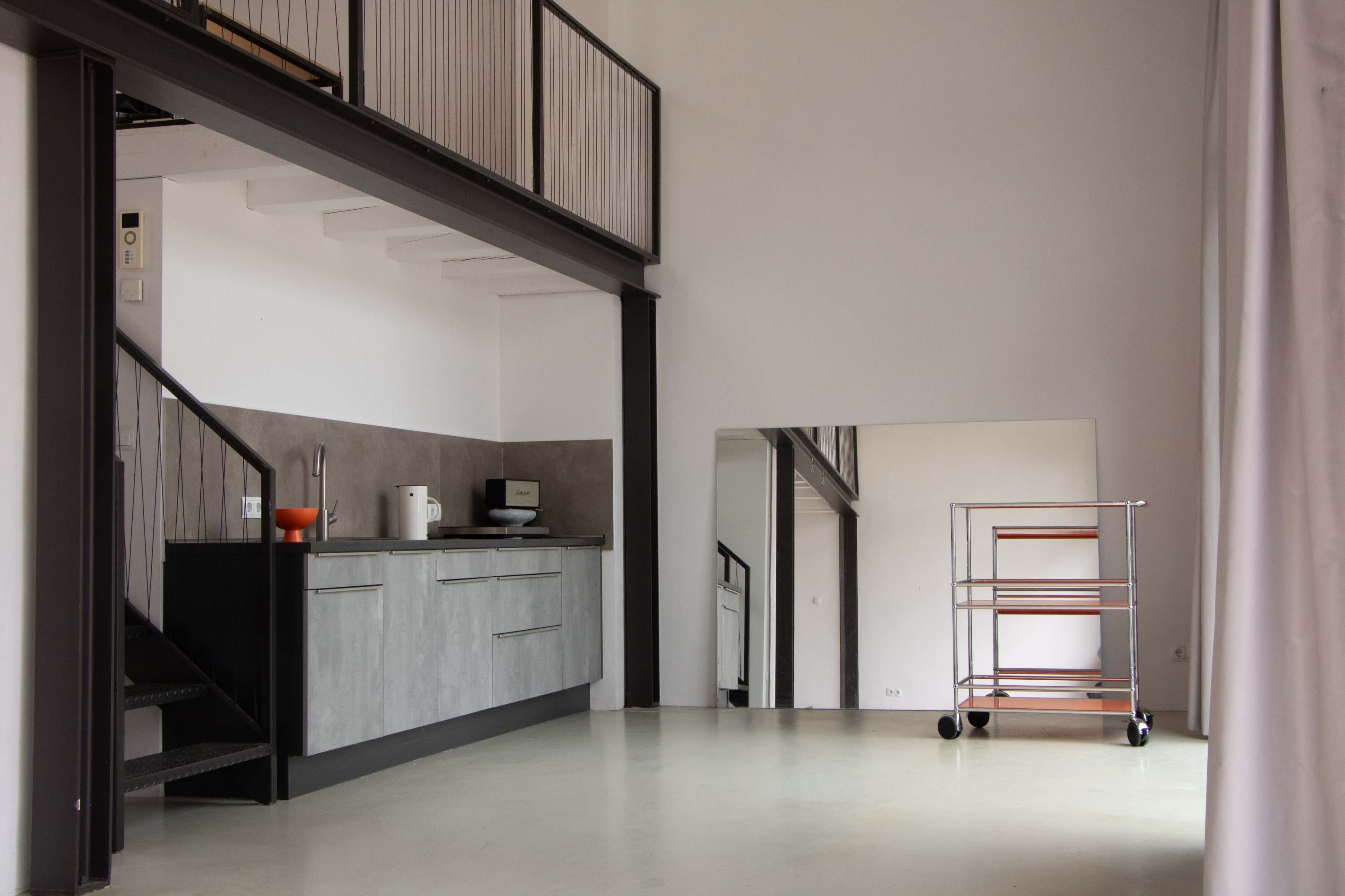 The image shows a minimalistic, contemporary kitchen area with a staircase and a large mirror reflecting part of the space.