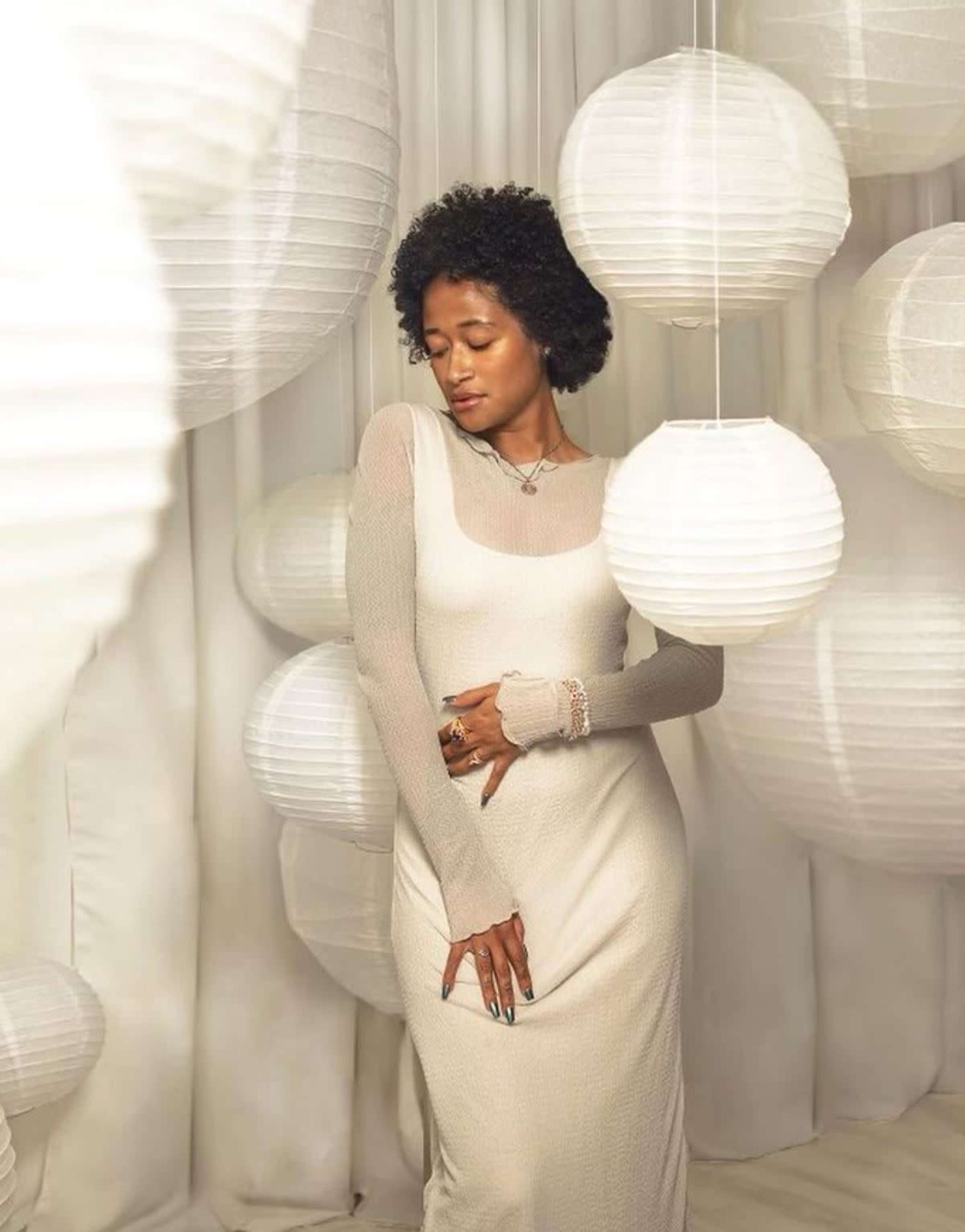 A woman in a long, fitted dress poses among white paper lanterns hanging in a softly lit space.