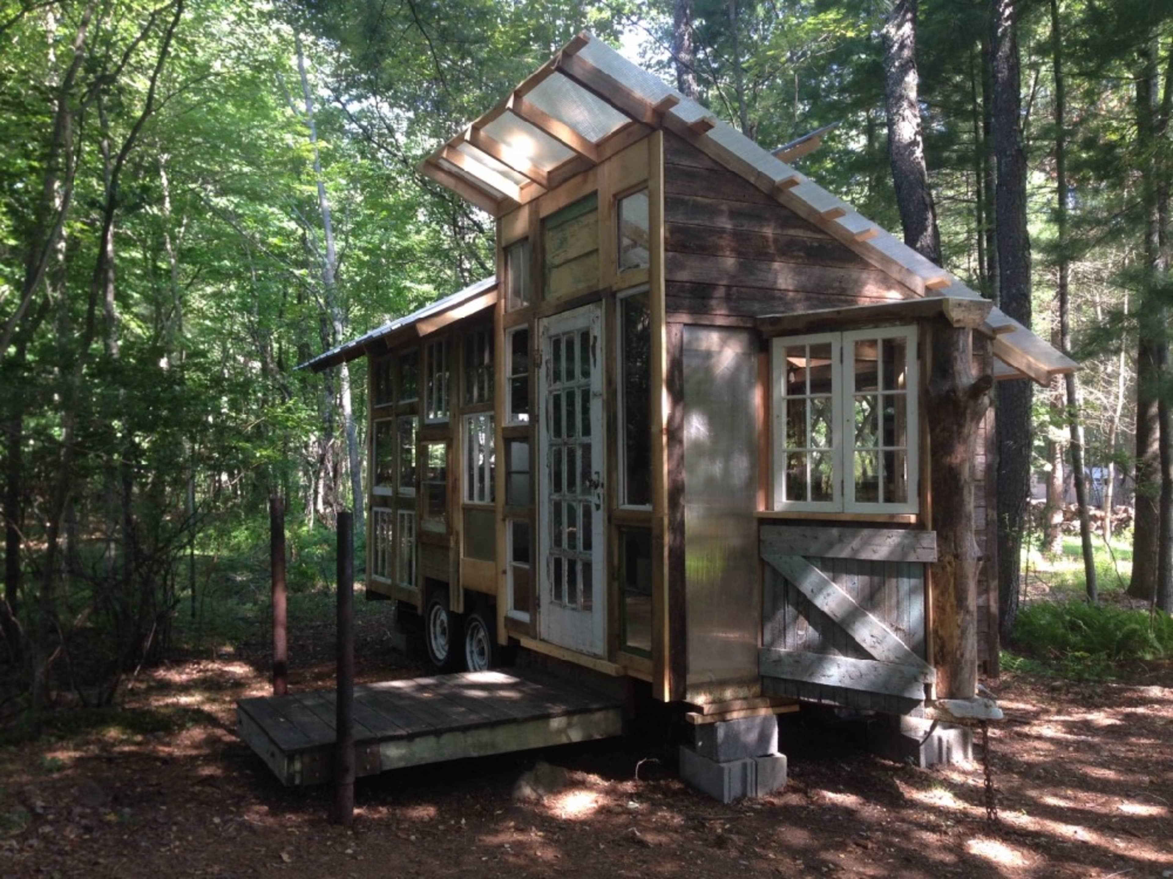 A small wooden house on wheels with multiple windows is situated in a wooded area.