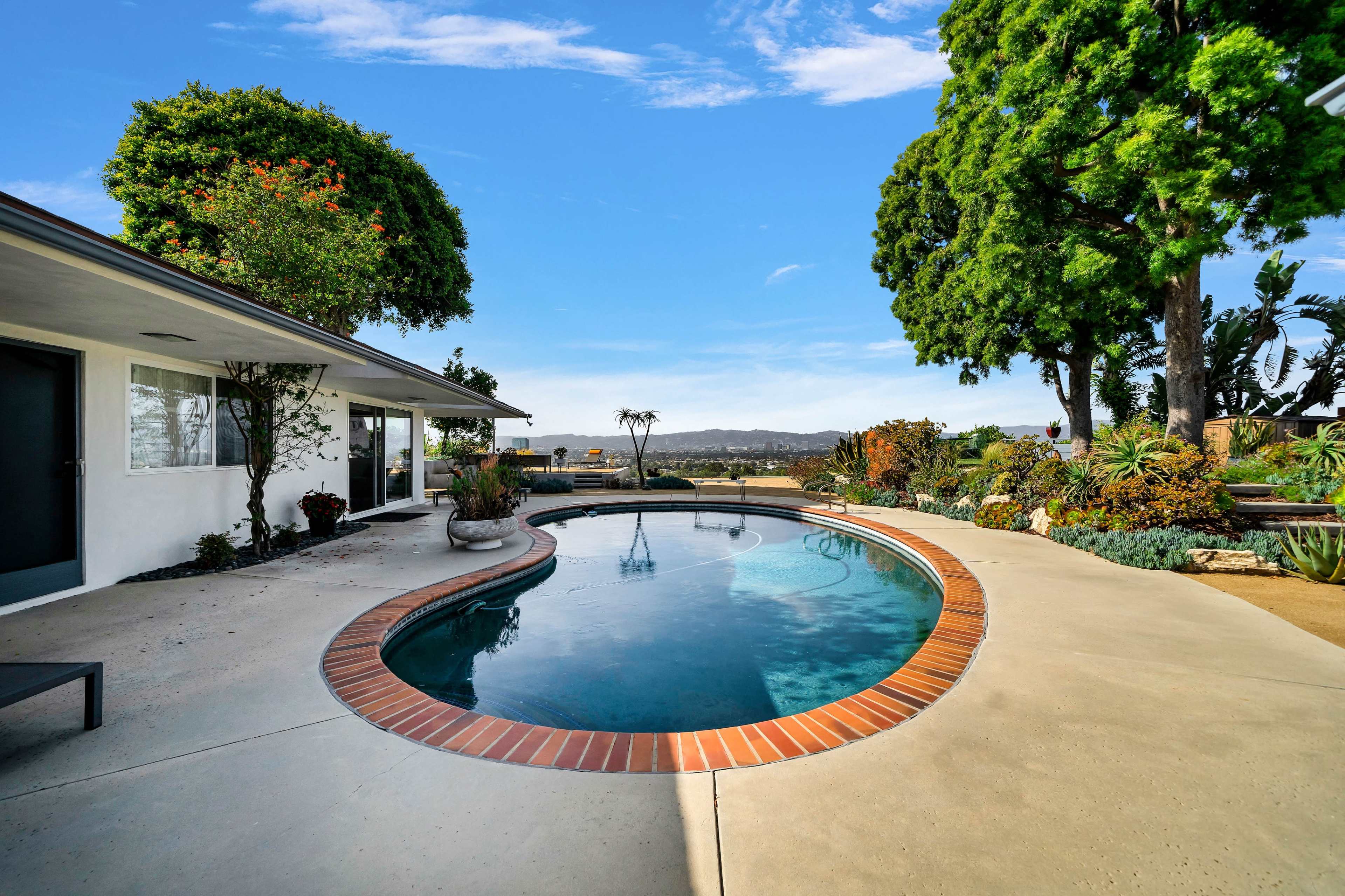 A curved pool with a brick edge is situated next to a modern house surrounded by landscaped gardens and trees, with a view of distant hills.