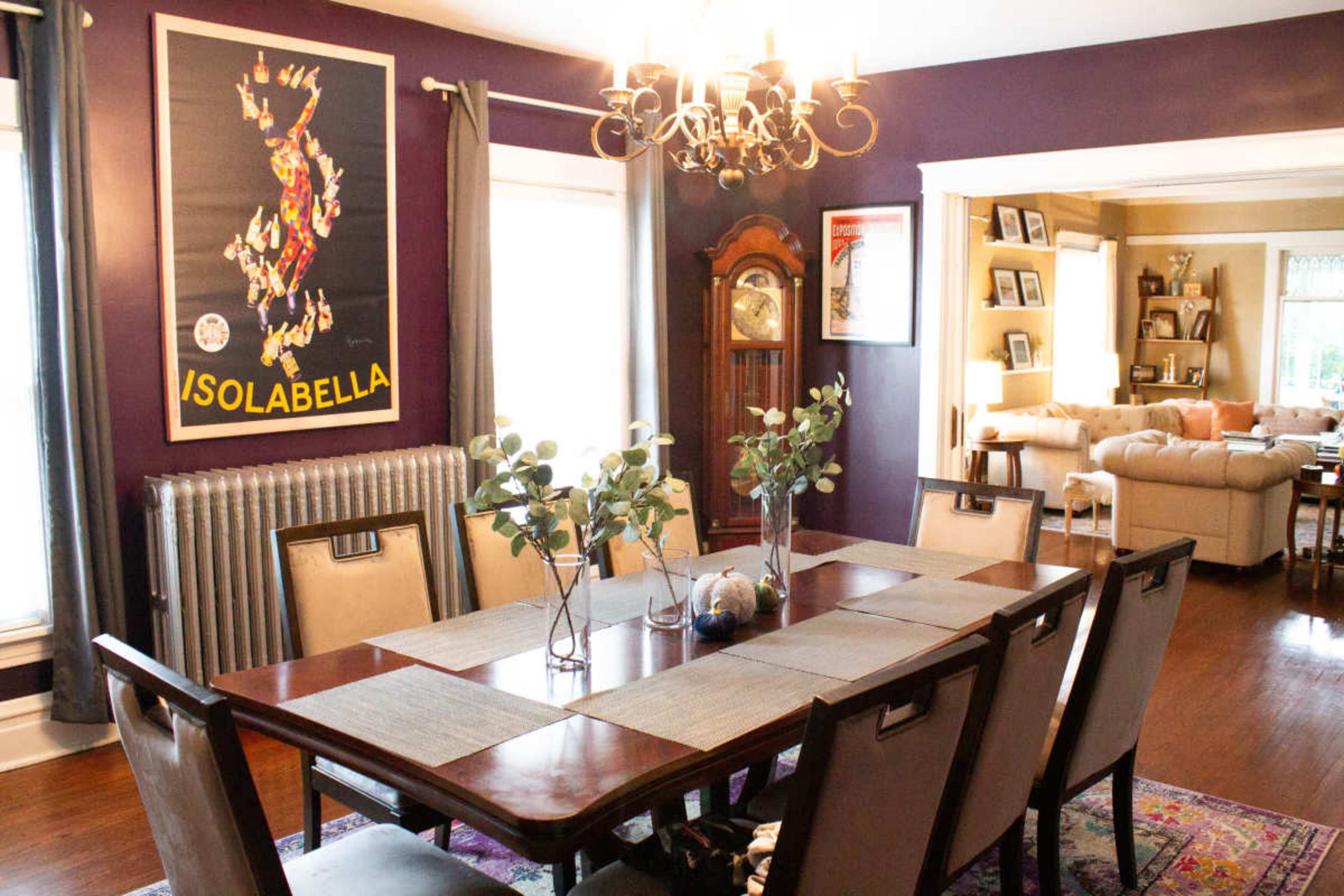 A dining room features a long wooden table with chairs surrounded by purple walls, a chandelier overhead, and a vintage poster on display.