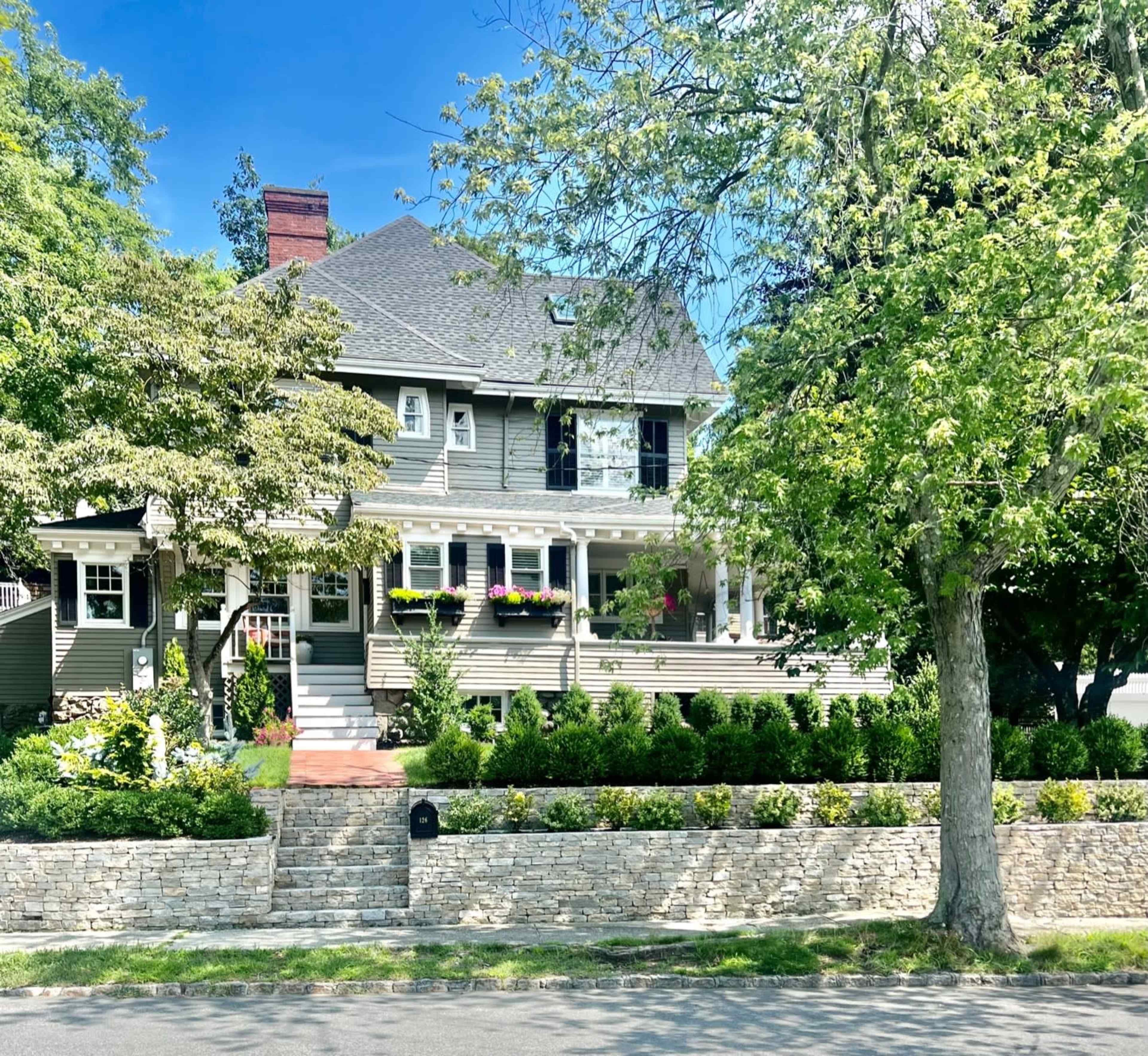 A large, two-story house with a gray exterior is surrounded by well-maintained shrubs and trees, featuring a front porch adorned with flower boxes.