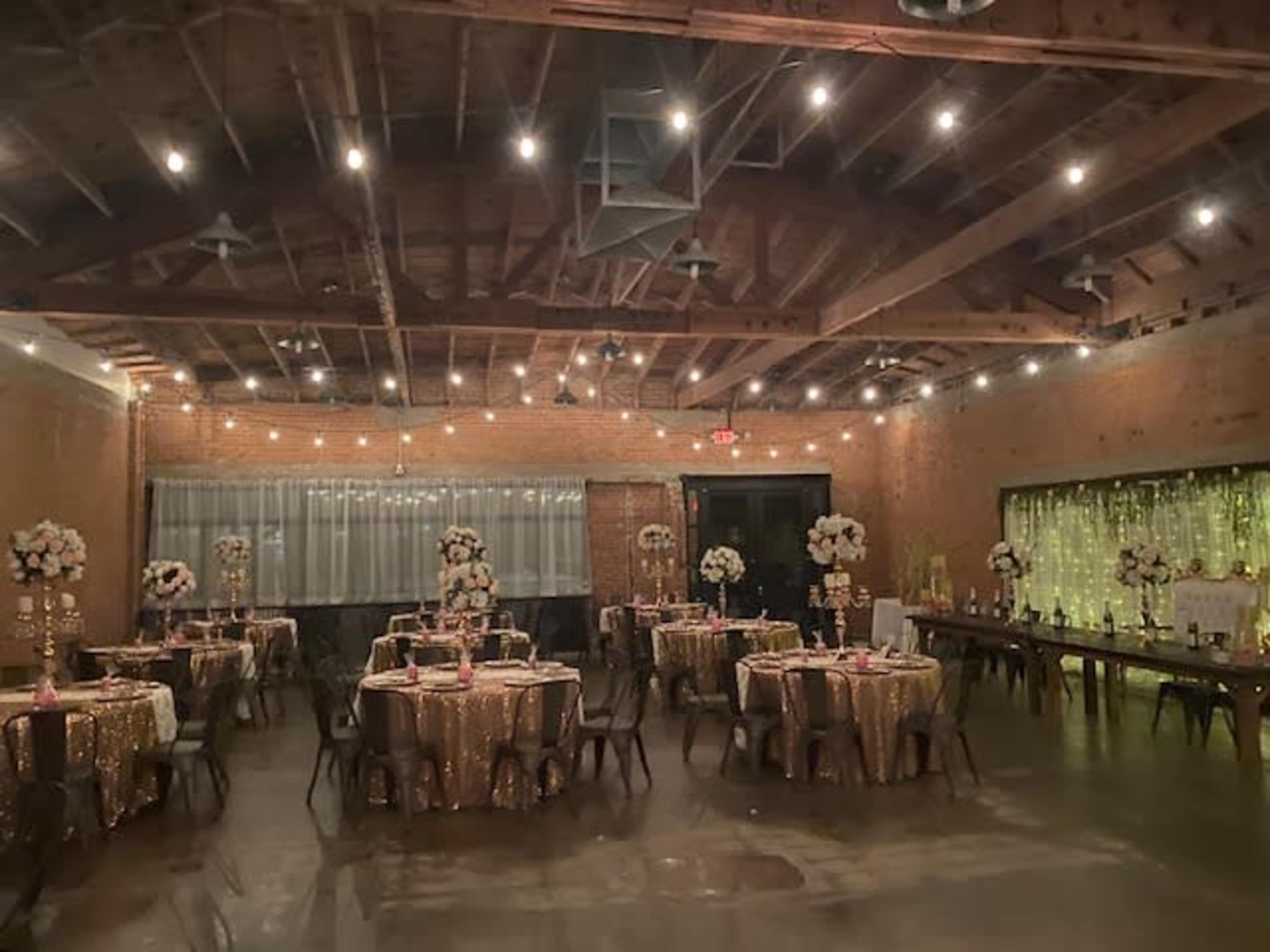A banquet hall with tables covered in gold tablecloths, adorned with floral centerpieces, and string lights creating a warm atmosphere.