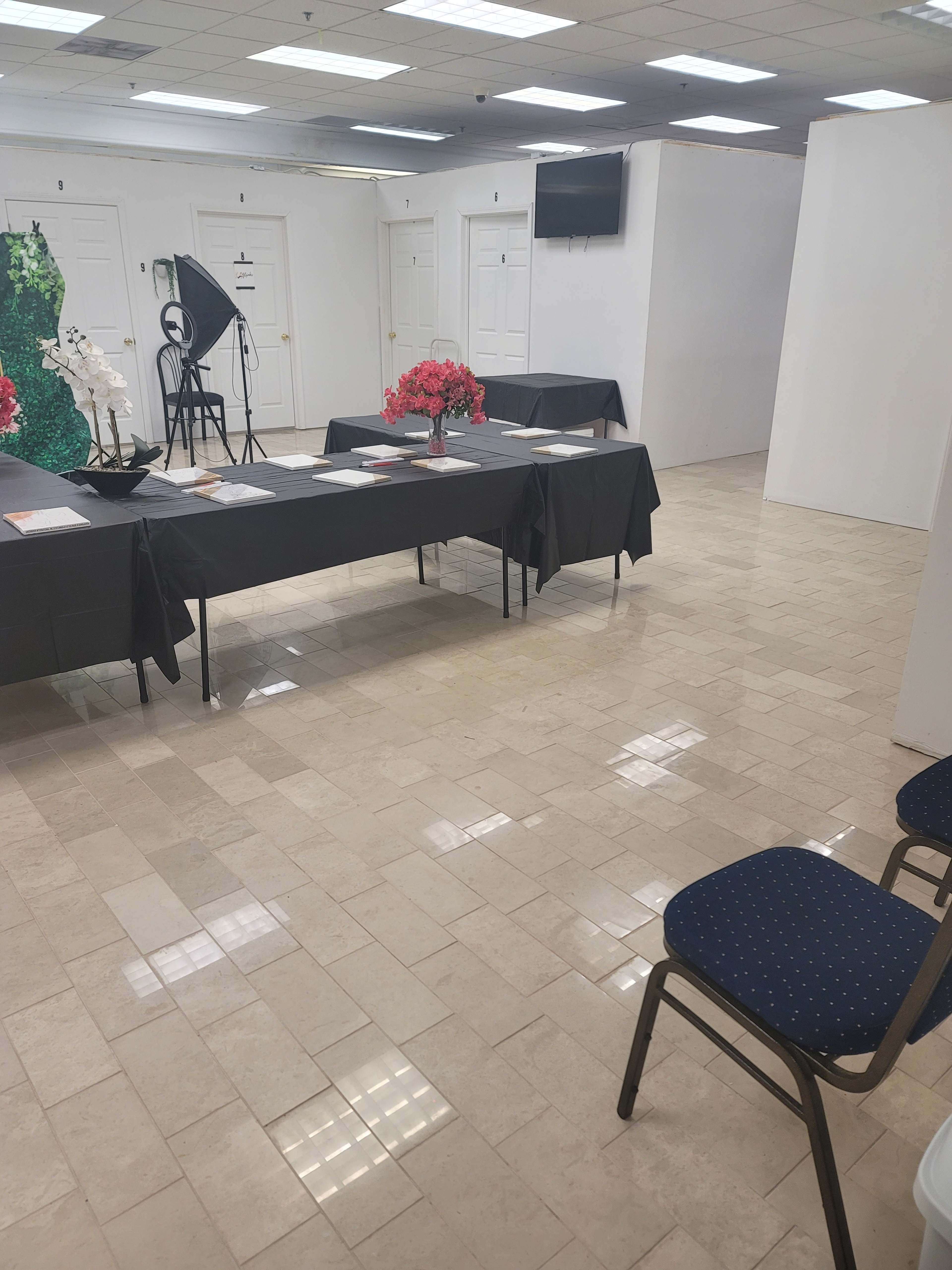 The image shows a large, well-lit room with tiled flooring, featuring several black tables set up with decorative flowers and chairs arranged nearby.