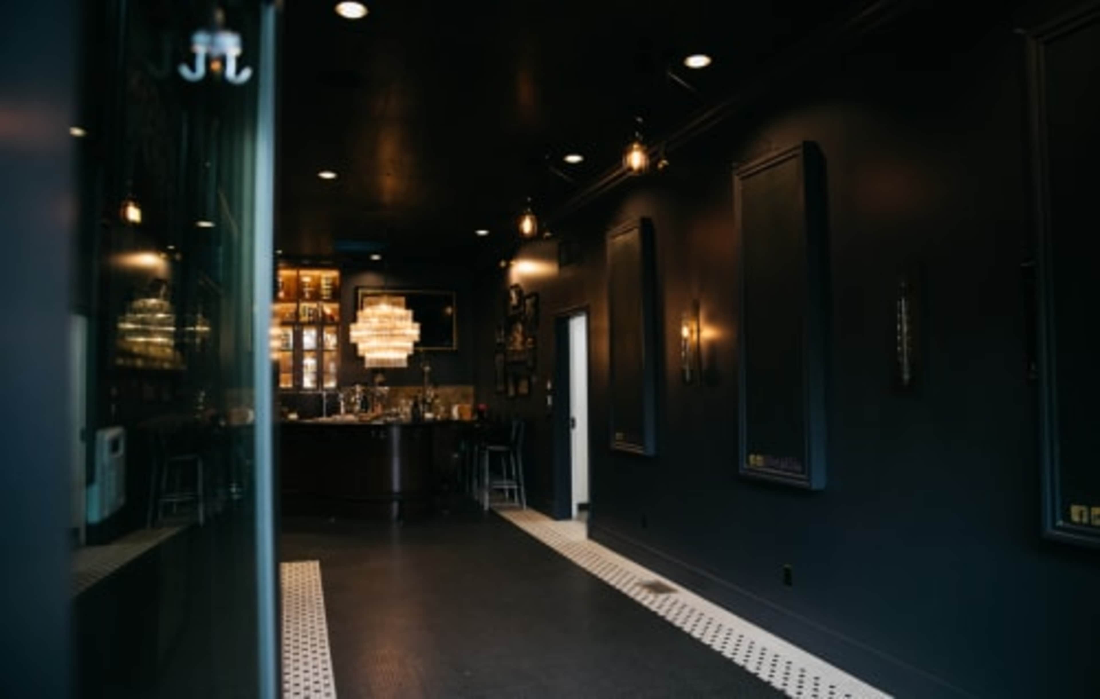 A narrow, dark-colored hallway leading to a bar area illuminated by pendant lights and featuring a large chandelier.