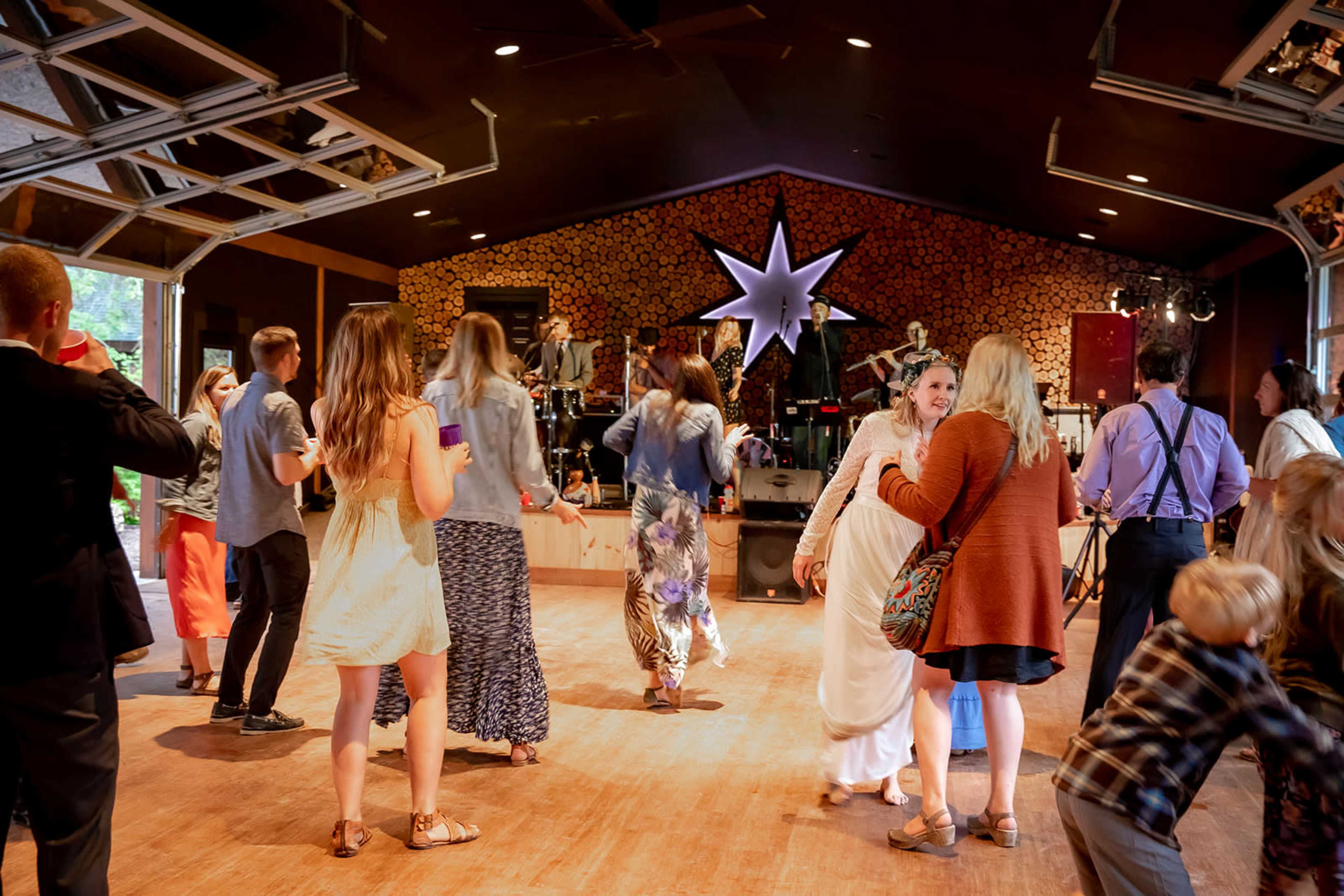 A group of people dance and socialize in a spacious indoor venue with a wooden floor and a band performing on stage.