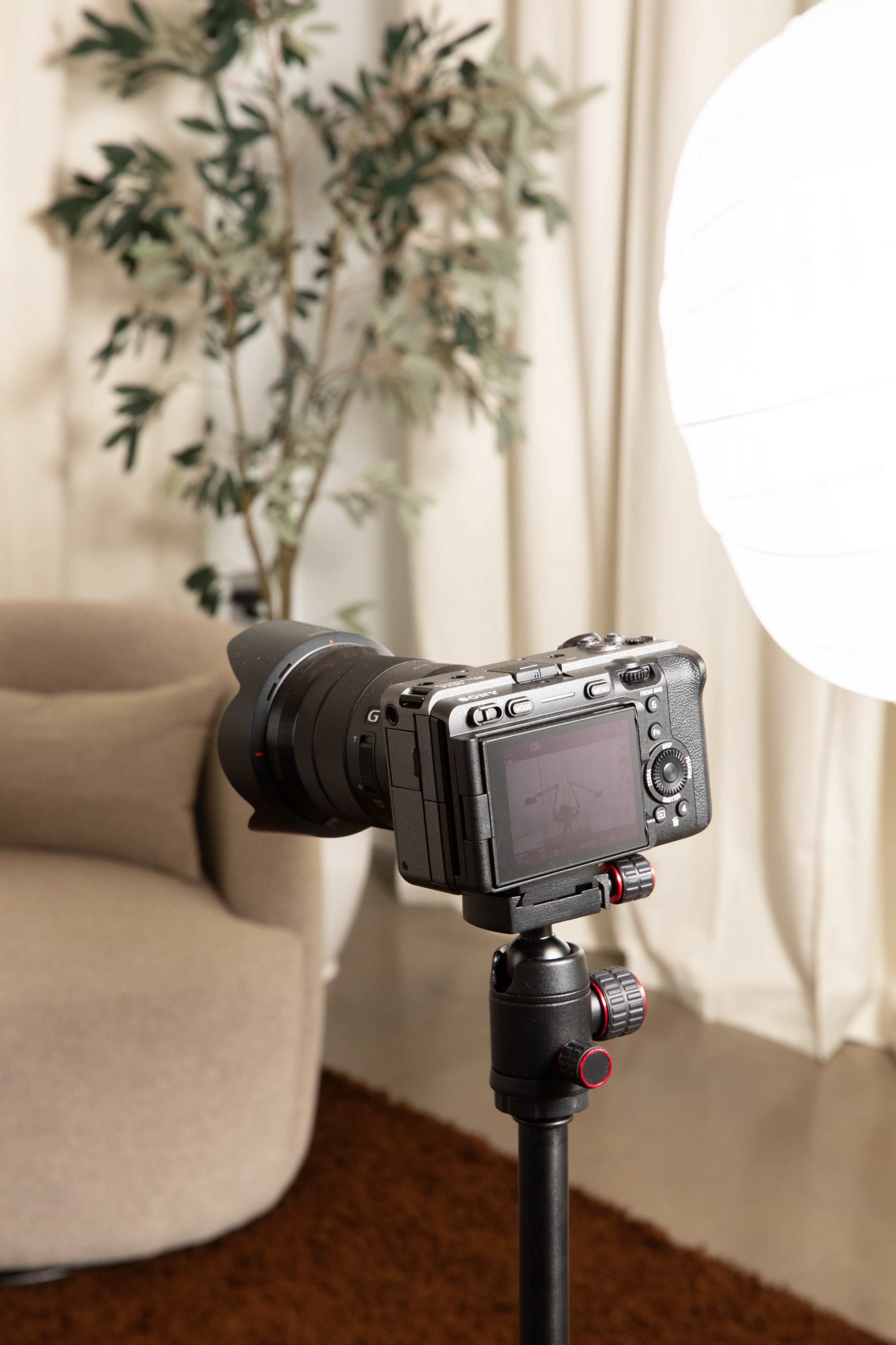 A camera is positioned on a tripod facing a soft light source with a large white lamp, near a beige sofa and a potted plant.