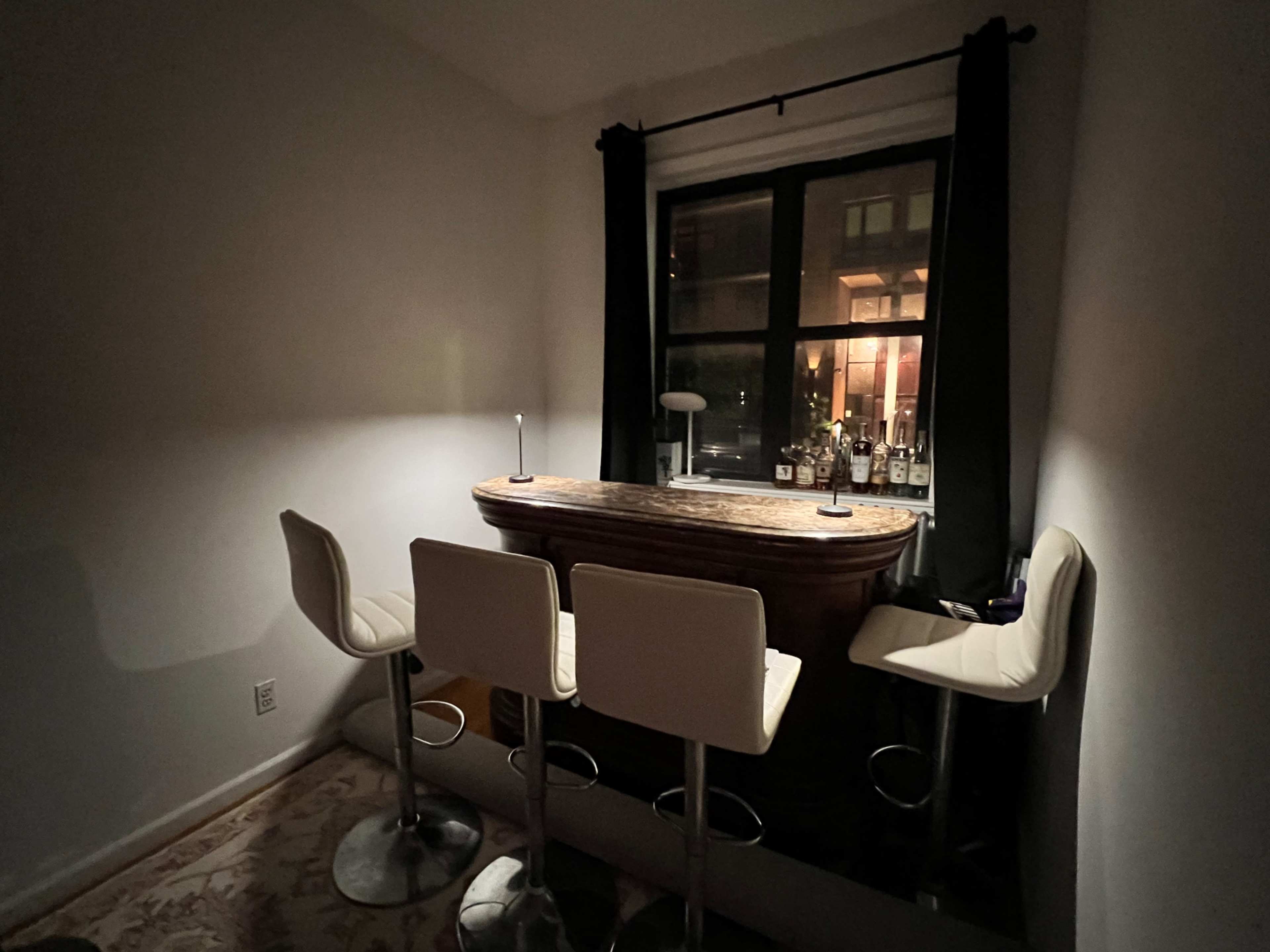 A small bar area with three white stools situated next to a window displaying bottles of liquor on the sill.