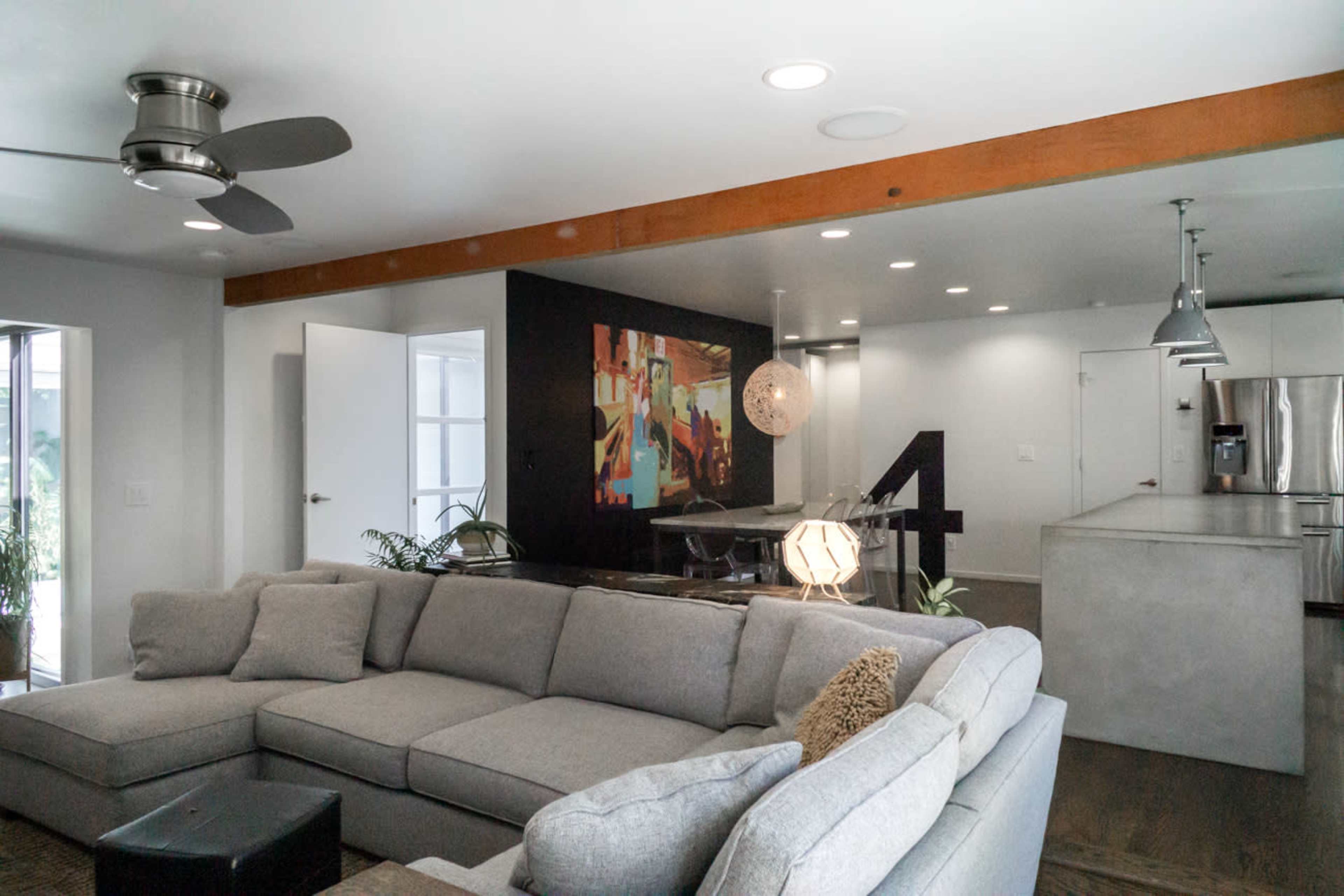 A modern living space featuring a gray sectional sofa, a dining area with a colorful wall mural, and a kitchen with stainless steel appliances.