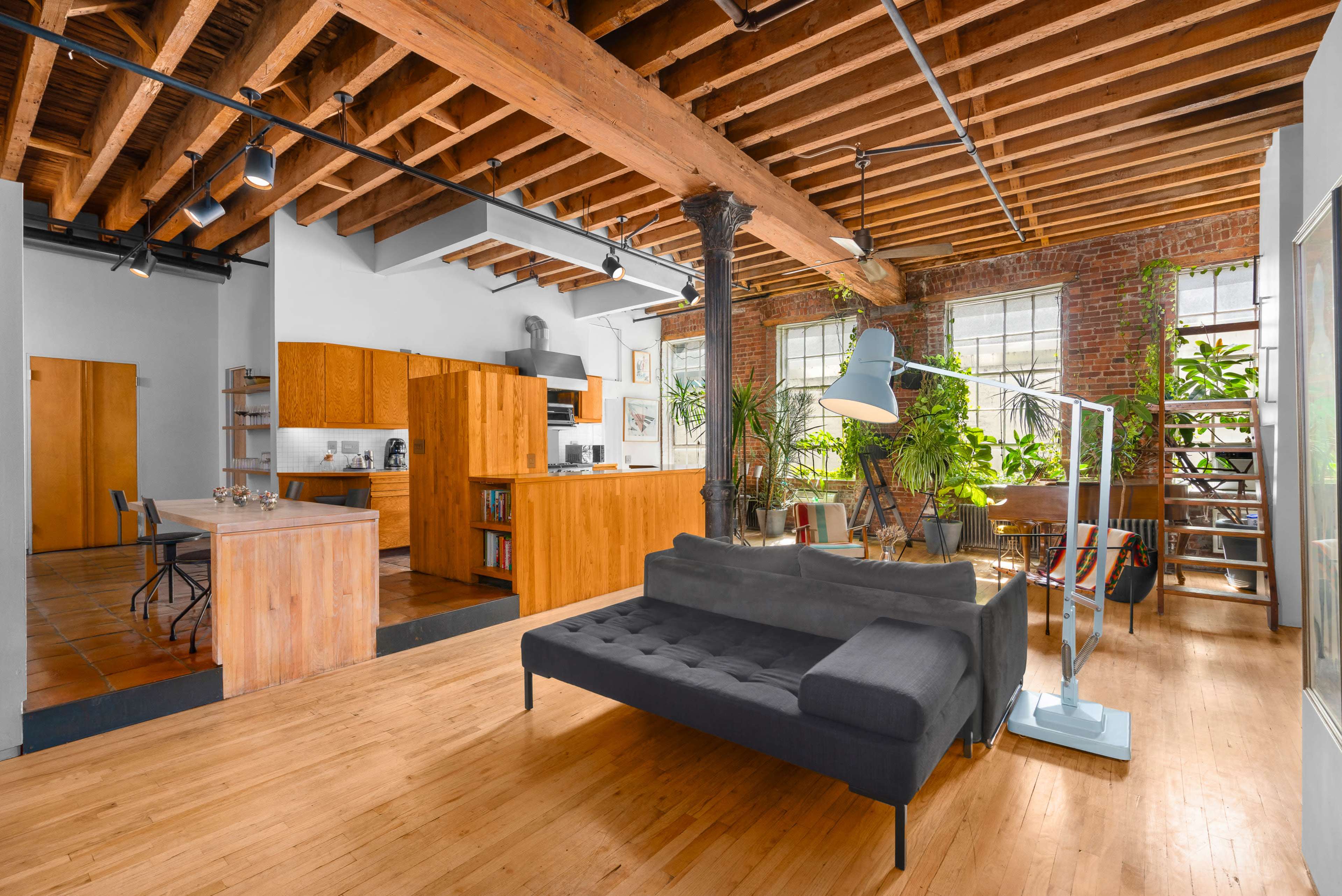 The image shows a spacious, modern living area with exposed wooden beams, a gray sectional couch, and a kitchen area featuring wooden cabinets and a dining space.