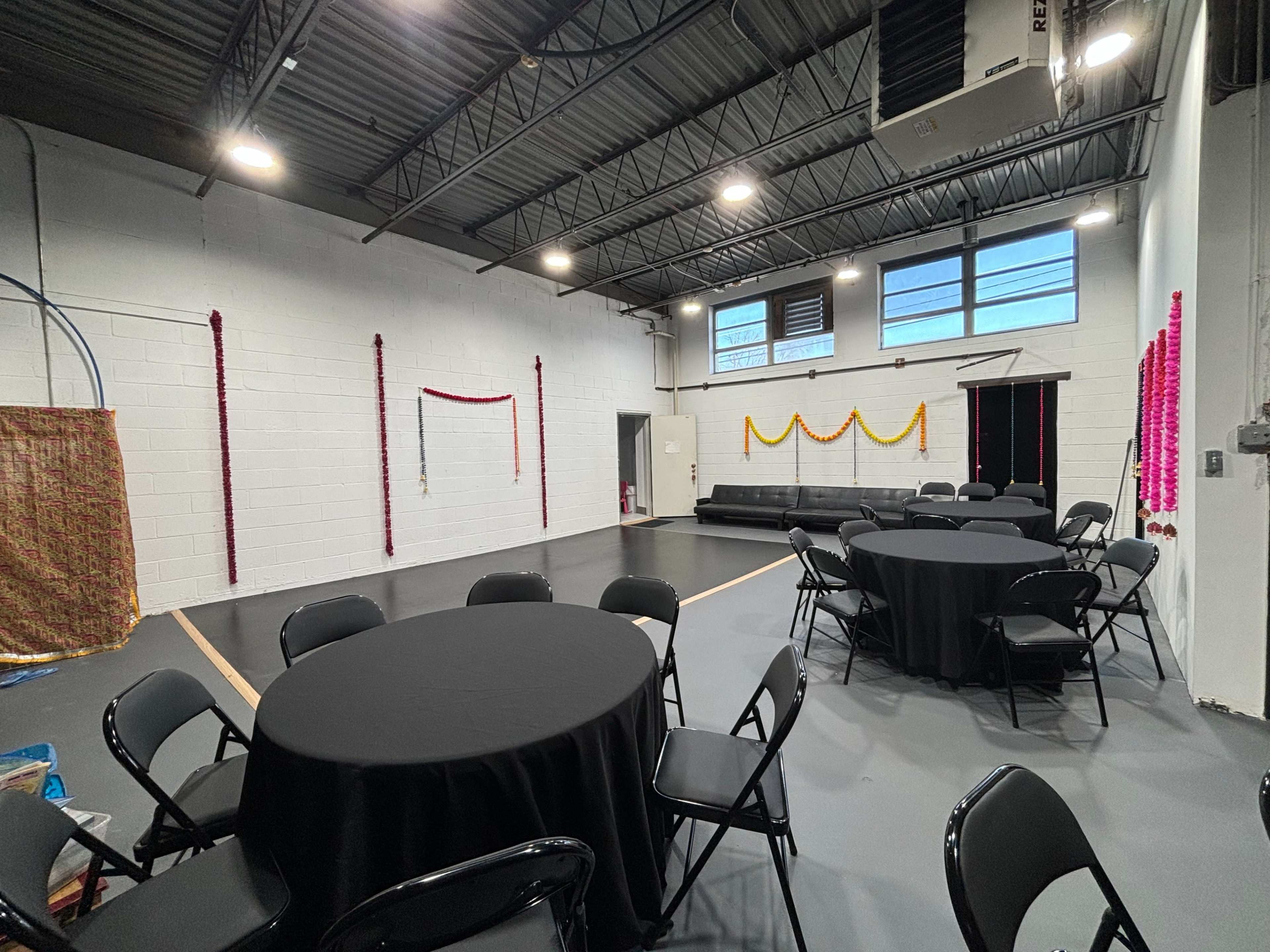 The image shows a spacious, well-lit indoor area with several round black tables and chairs arranged, and colorful decorations hanging on the walls.