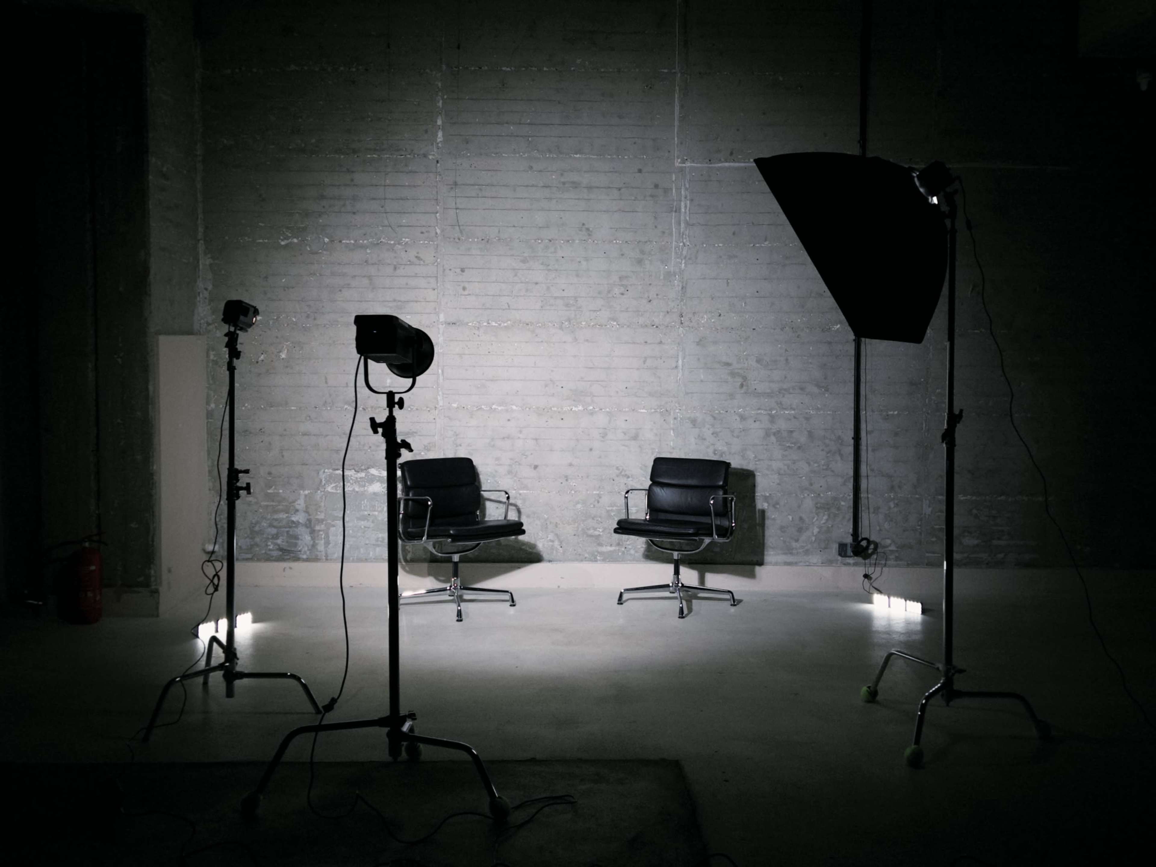 The image shows two black chairs under soft lighting in a minimalistic studio setup with photography equipment positioned around them.