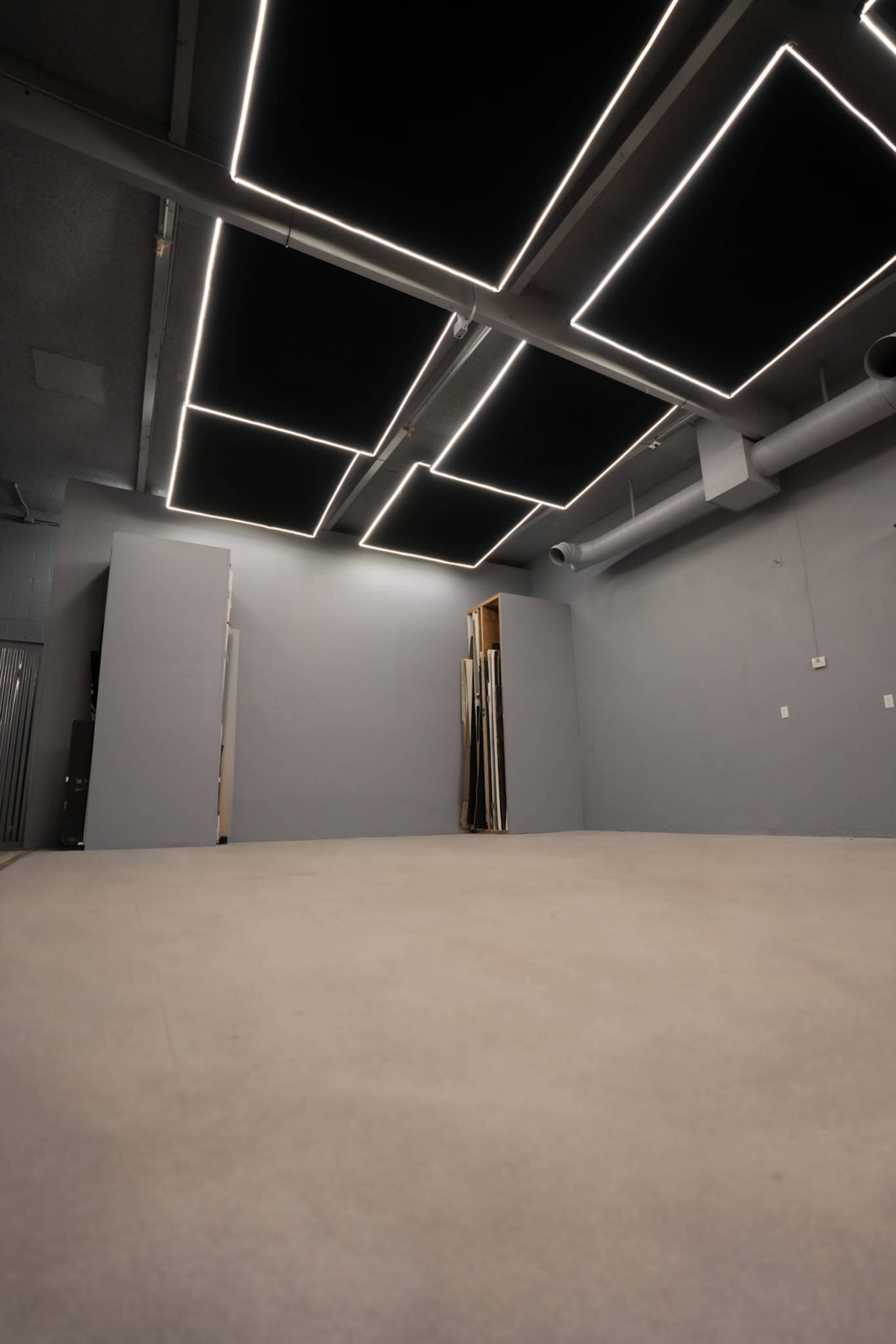 The image shows an empty room with a concrete floor and modern black ceiling panels illuminated by rectangular LED lights.