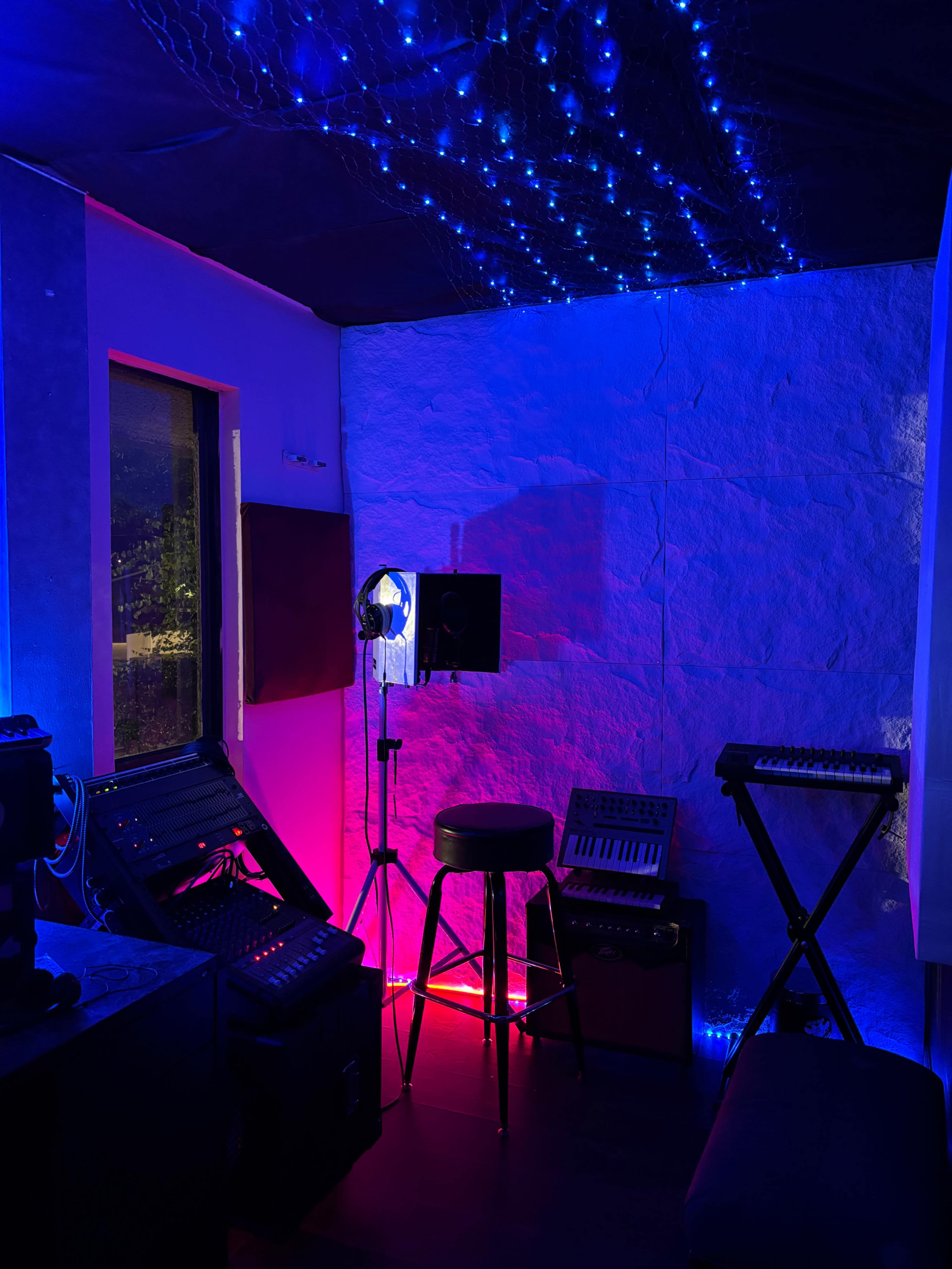 The image shows a small room with colored LED lights, a music stand, a stool, and various musical equipment against the walls.
