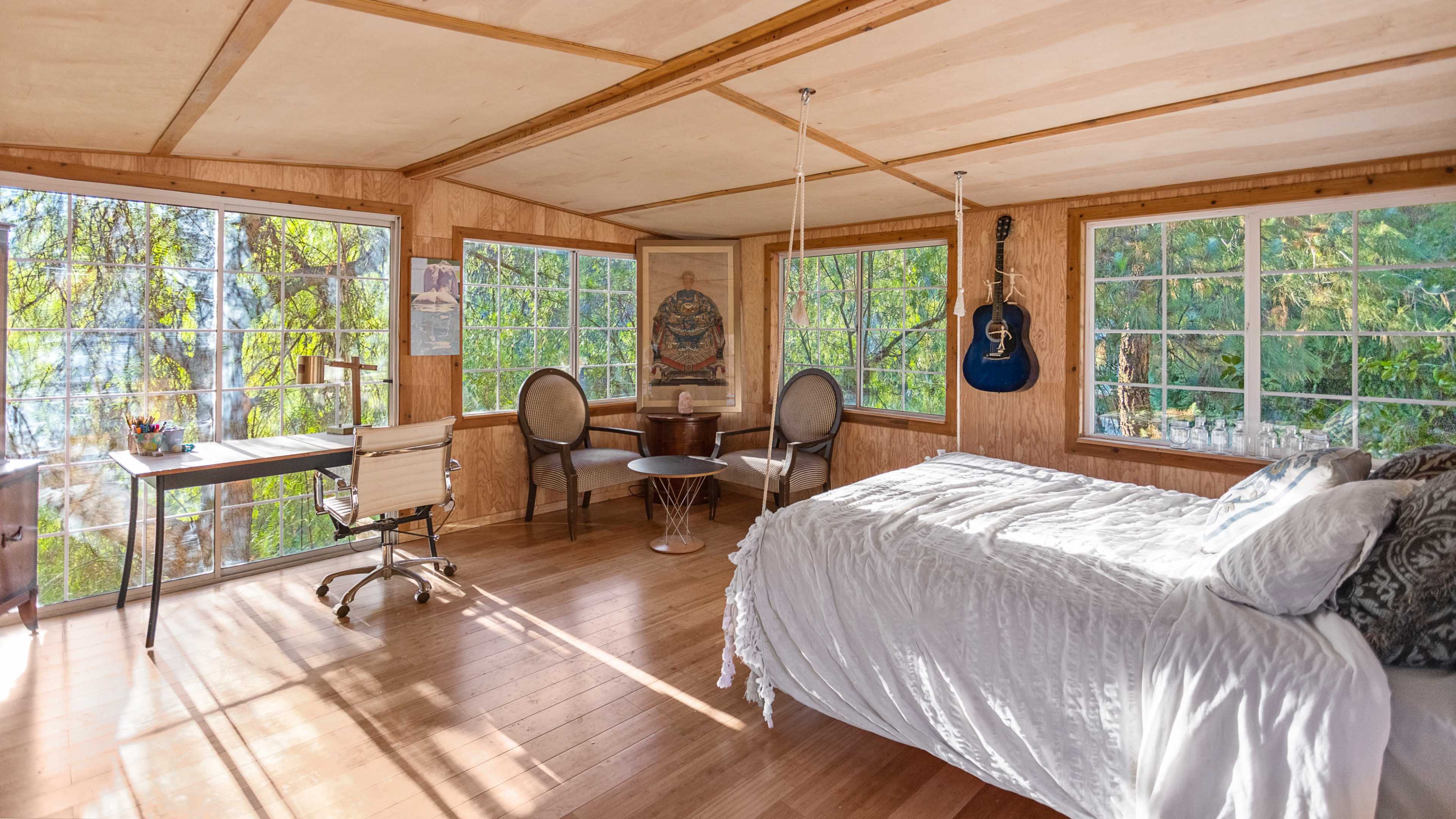 The room features a wooden interior with large windows, a bed, a desk, and a guitar hanging on the wall, overlooking a natural landscape.