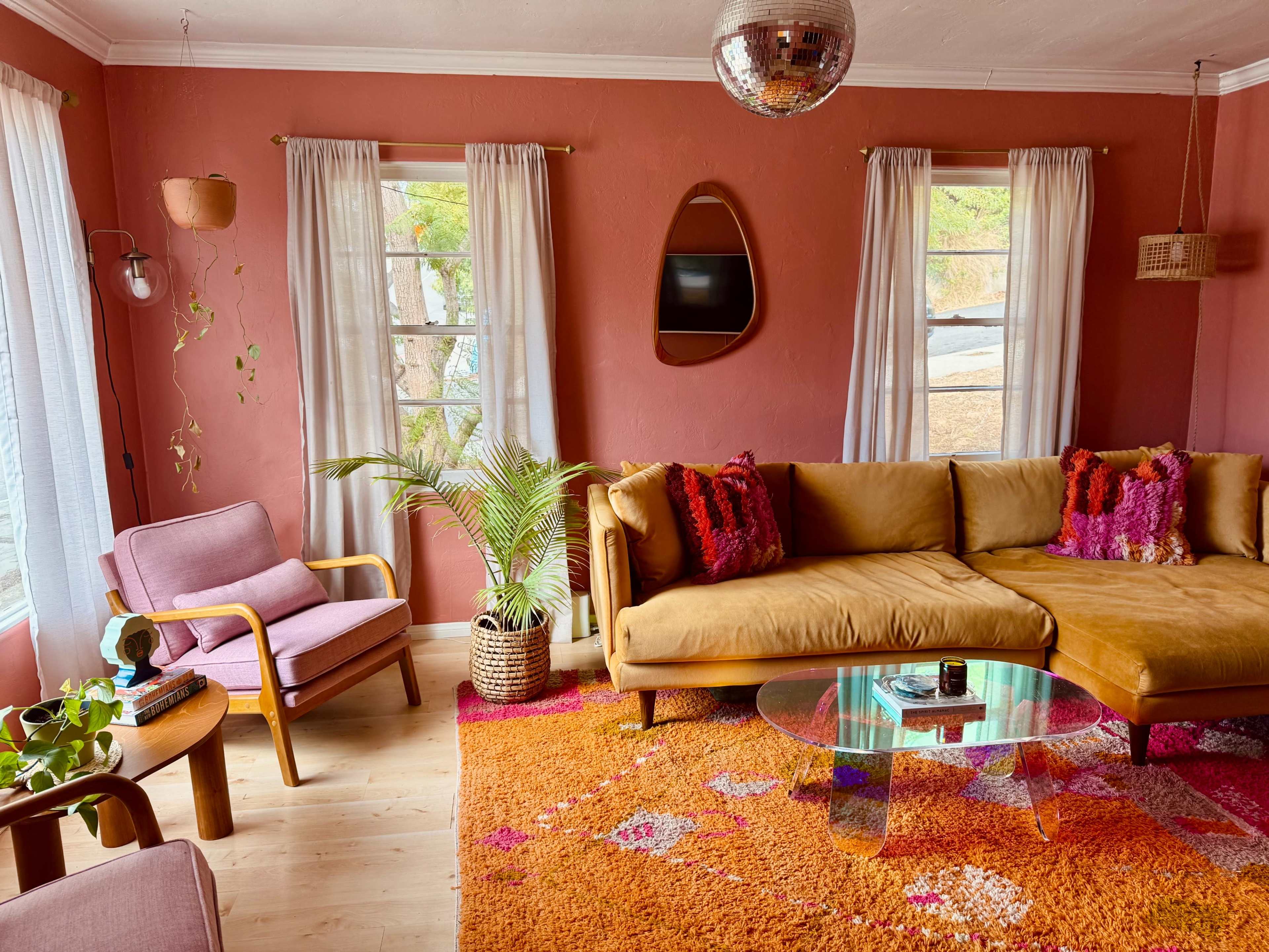A cozy living room features a brown sectional sofa, a pink armchair, a glass coffee table, and colorful rugs, with sunlight streaming through the windows.